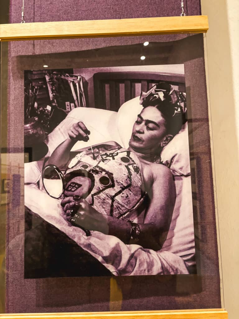 Black and white photograph of Frida Kahlo painting on her medical cast while lying in bed, displayed inside the Frida Kahlo Museum.