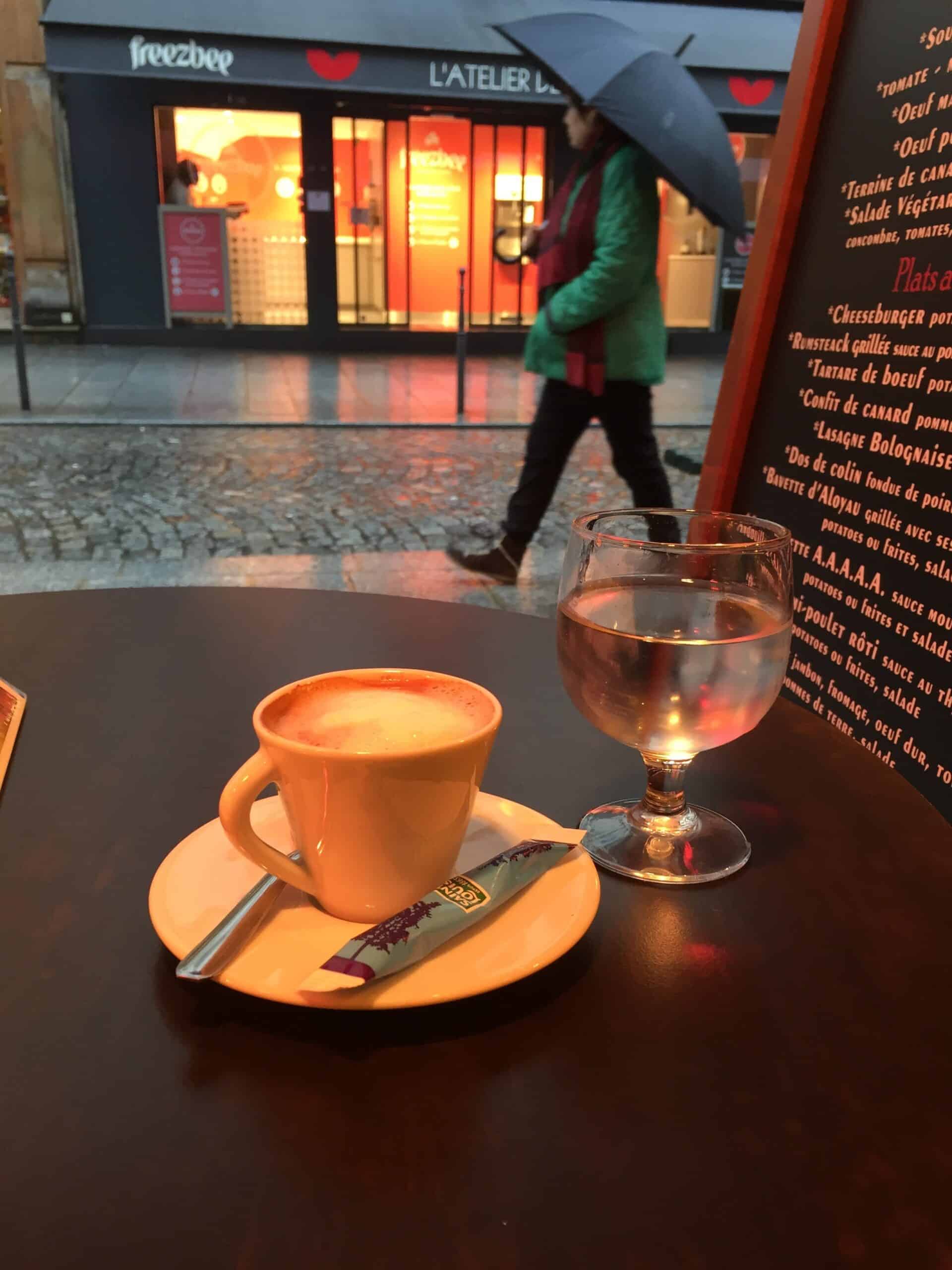 Warm-lit espresso and glass of water on a rainy evening in Paris, seen from a cozy café window.