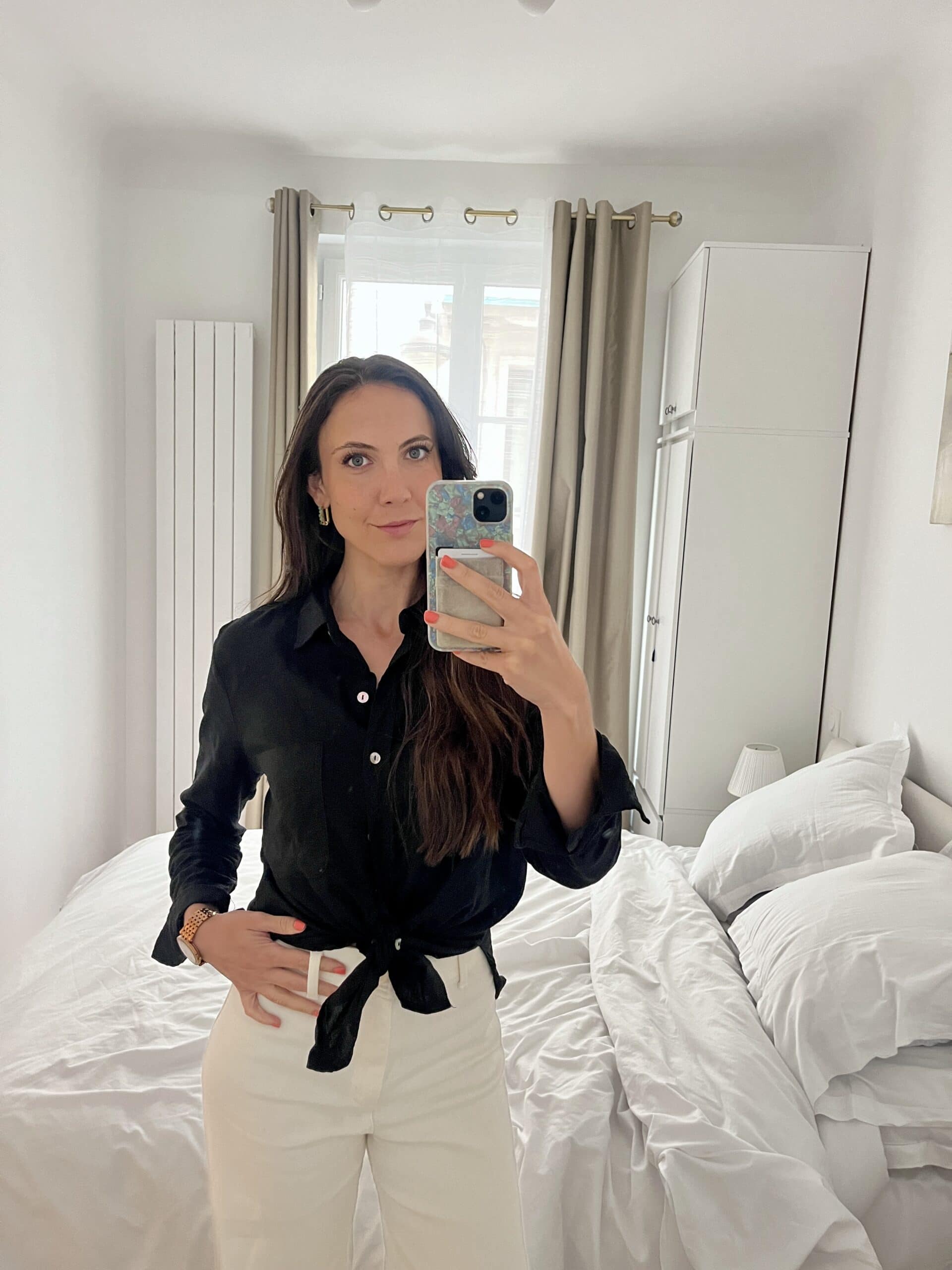 Woman in a black blouse and white pants taking a mirror selfie in a bright Paris apartment, showing what to wear for 3 days in Paris.