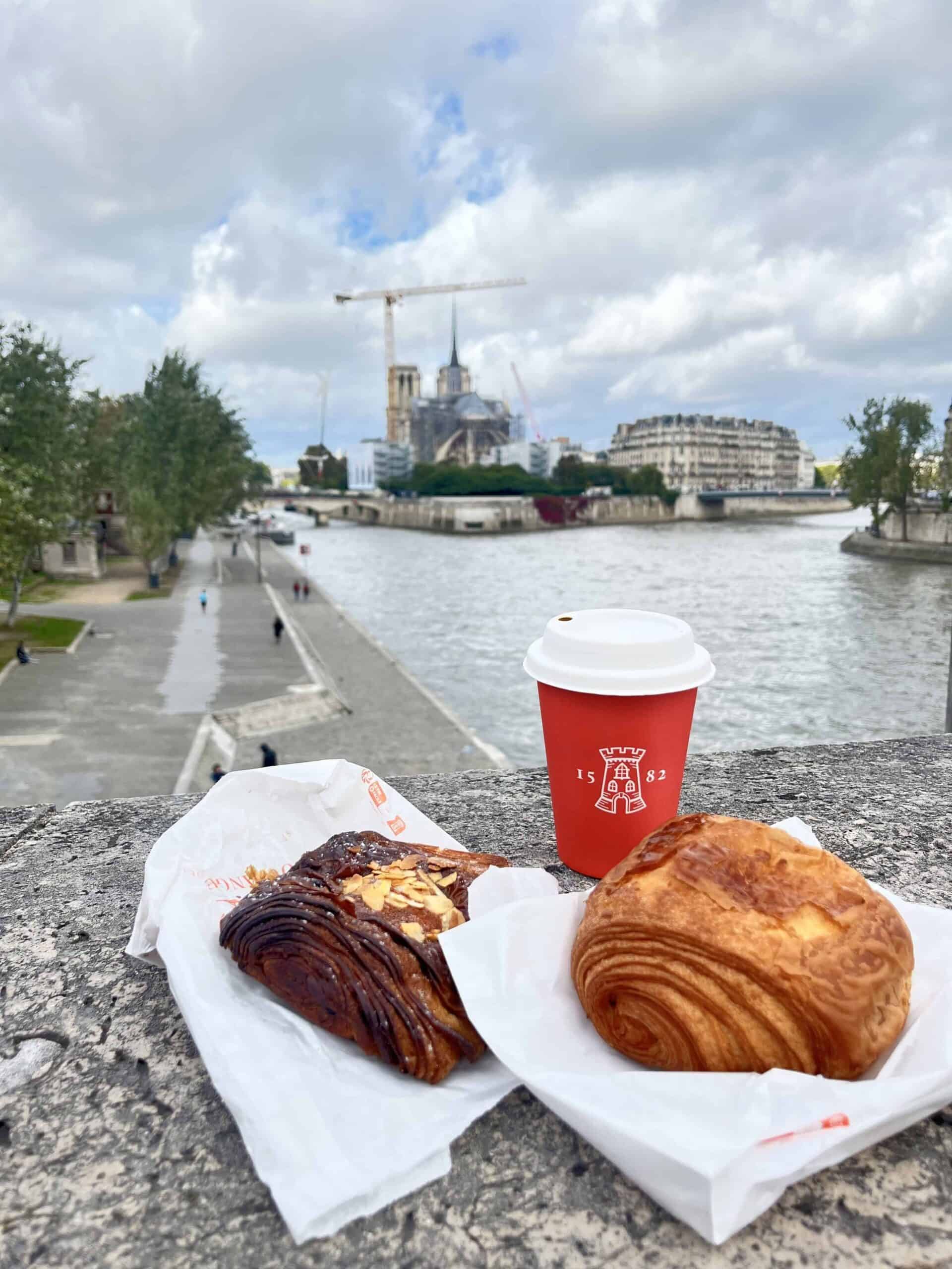 Breakfast on a stone ledge with pain au chocolat, almond croissant, and coffee overlooking the Seine and Notre-Dame—classic 3 days in Paris moment.