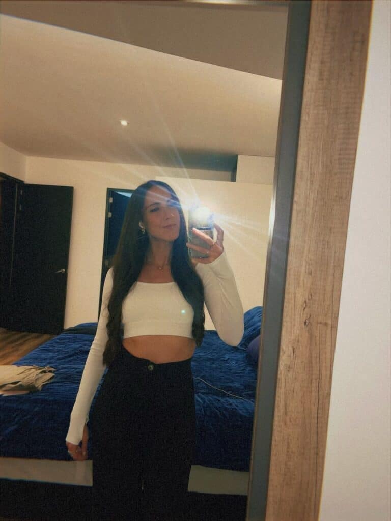 Woman taking a mirror selfie in a modern hotel room, wearing a white long-sleeve crop top and dark bottoms, showing stylish layered clothing perfect for Mexico City's variable temperatures