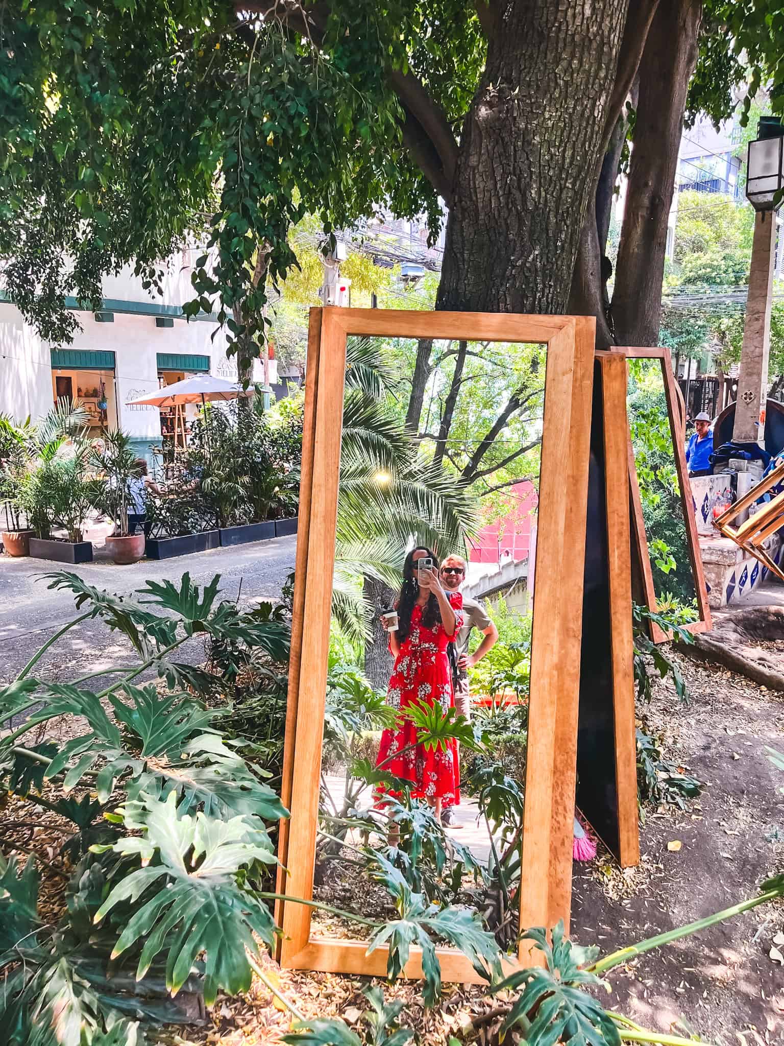 Woman in red floral dress posing in front of a large wooden mirror frame in a lush garden setting in Mexico City