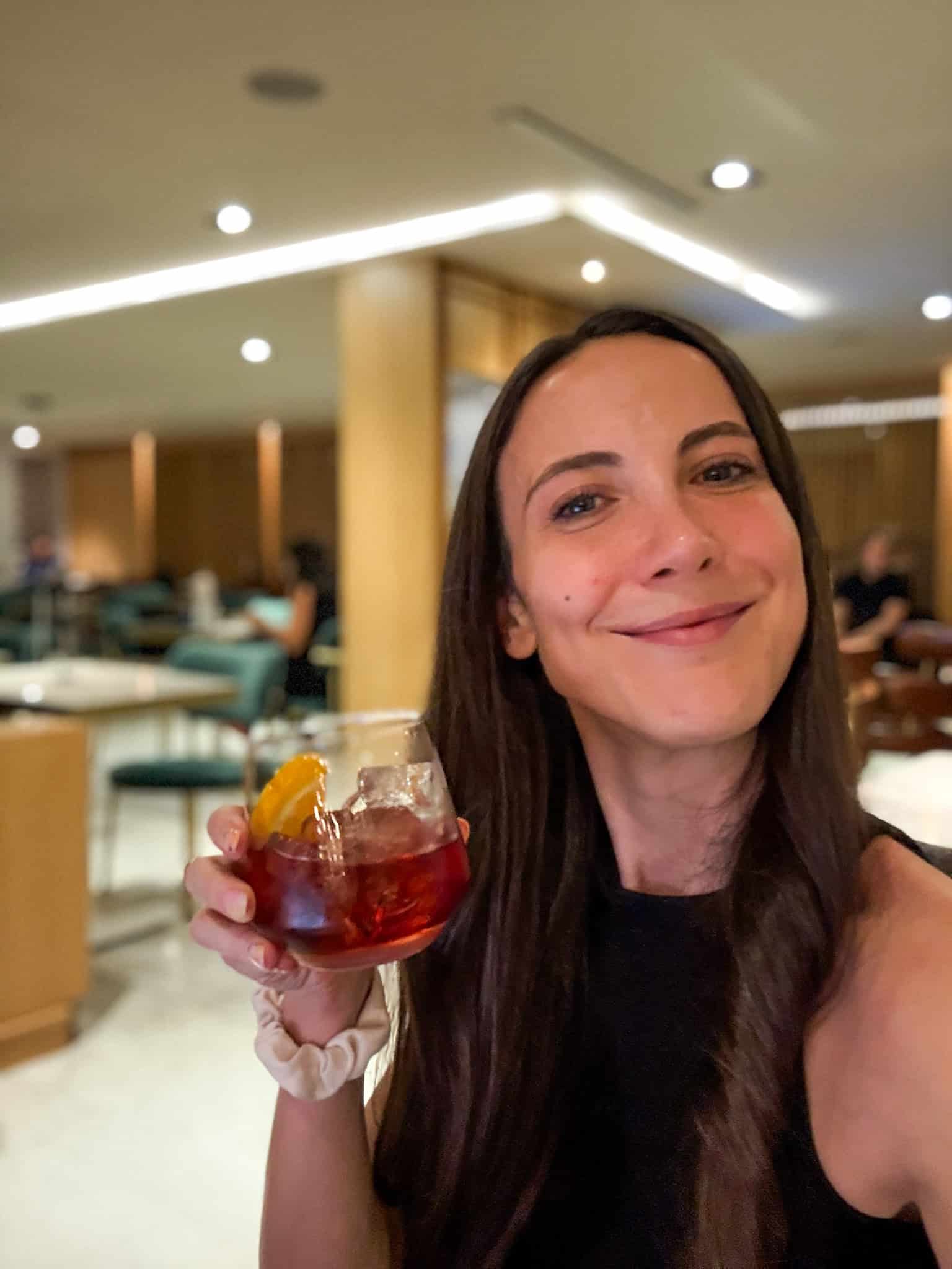 Woman holding a Negroni cocktail with orange garnish inside the Amex Centurion lounge Mexico City, smiling at camera