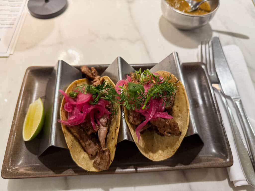 Rib eye tacos garnished with pickled red onions and fresh herbs, served on metal taco holders with lime wedge