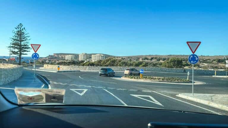 Driver's perspective approaching a roundabout while driving in Malta, showing typical road signs and coastal landscape views