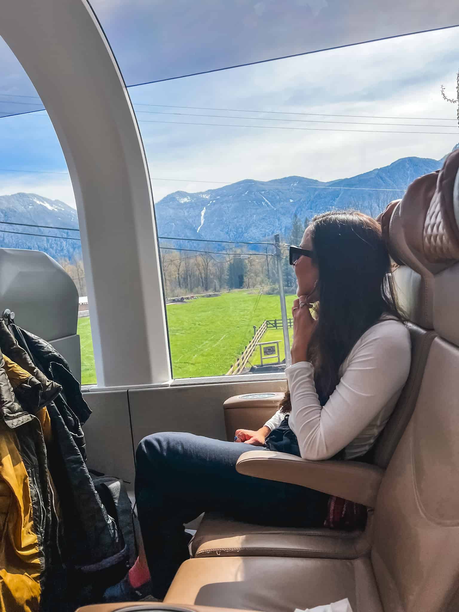 Passenger enjoying mountain views from Rocky Mountaineer train GoldLeaf coach window overlooking snowy peaks and green valleys in British Columbia