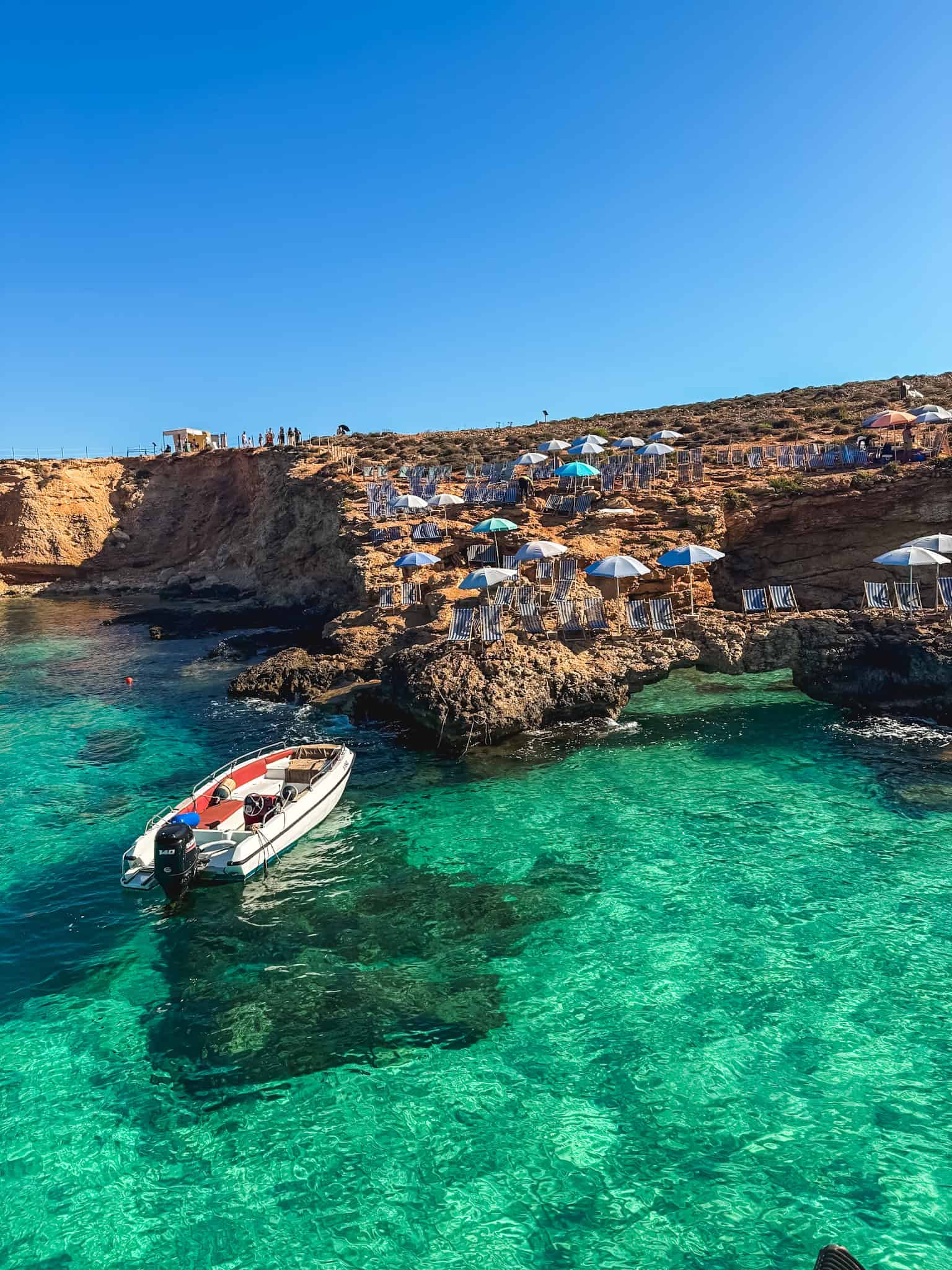 Blue Lagoon Comino with crystal clear turquoise waters, boat, and sunbathers on rocky cliffs - best Malta beaches