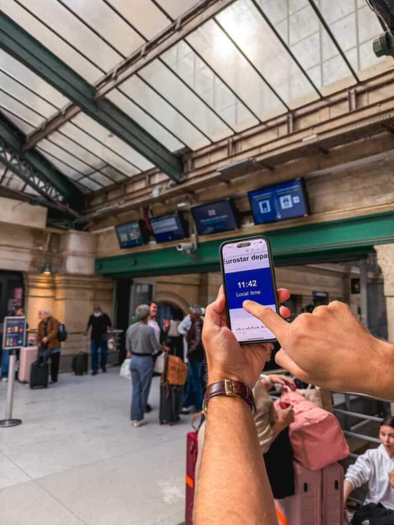 Close-up of a smartphone displaying the Eurostar departure time inside the Gare du Nord terminal with travelers and departure boards in the background