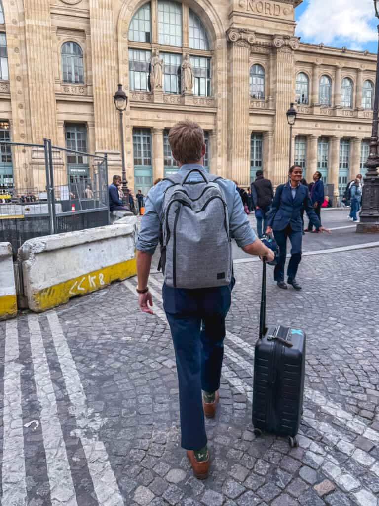 A solo traveler with a backpack and rolling suitcase walking across Paris cobblestones toward Gare du Nord to board the train from paris to london