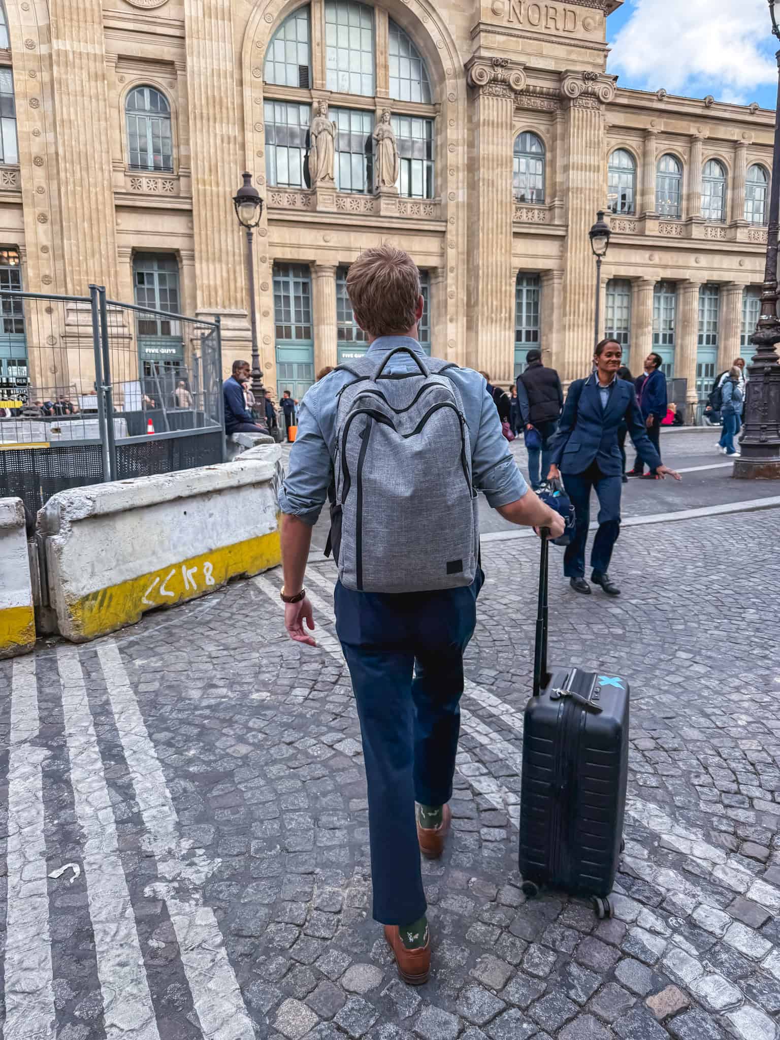 A solo traveler with a backpack and rolling suitcase walking across Paris cobblestones toward Gare du Nord to board the train from paris to london