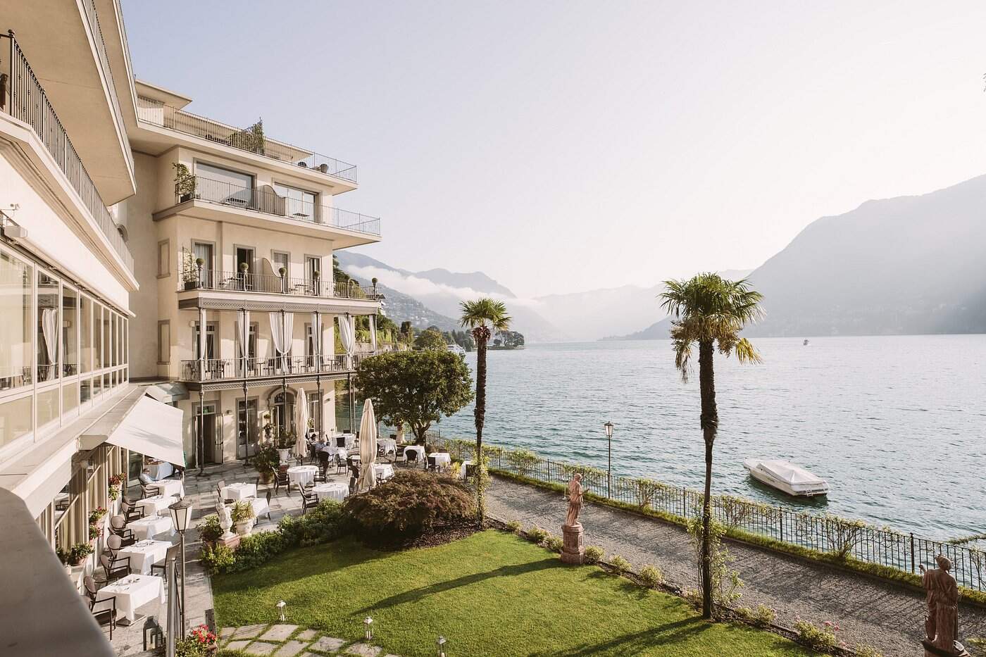 Elegant lakeside hotel terrace on Lake Como with alfresco dining, palm trees and misty mountain backdrop — luxury Italy summer travel