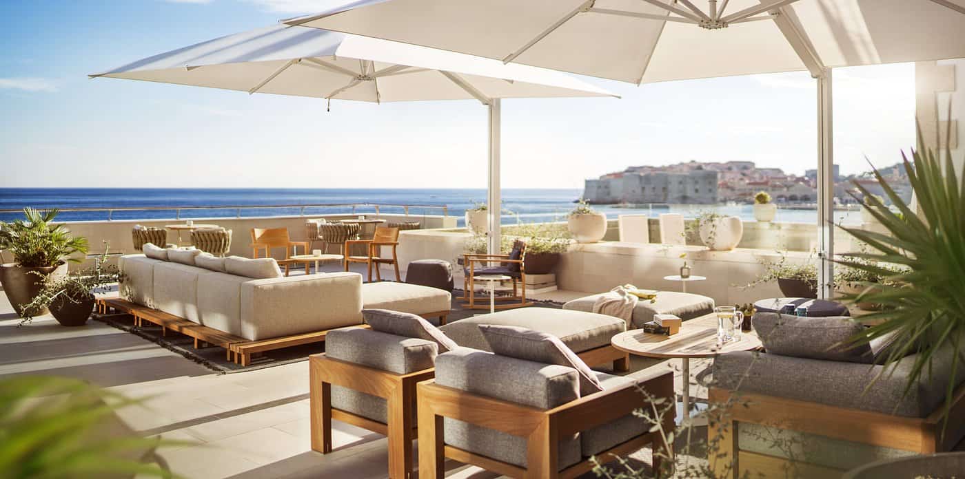 Upscale hotel rooftop lounge with neutral teak furniture overlooking the Adriatic Sea and Dubrovnik Old Town walls — European summer vacations in Croatia