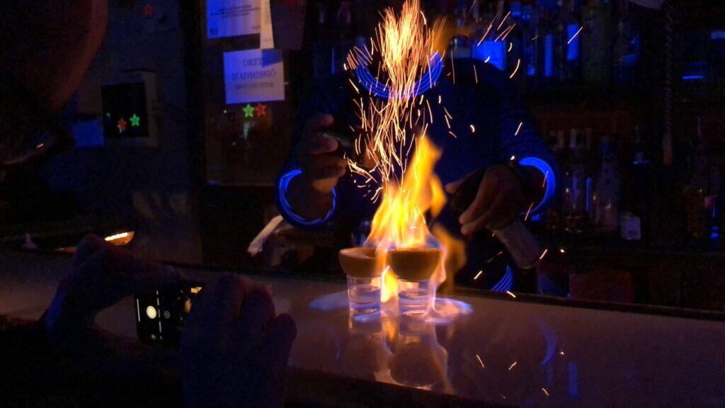 Bartender flaming shots at a Barcelona cocktail bar with dramatic fire and sparks under blue neon lights — Barcelona nightlife bars