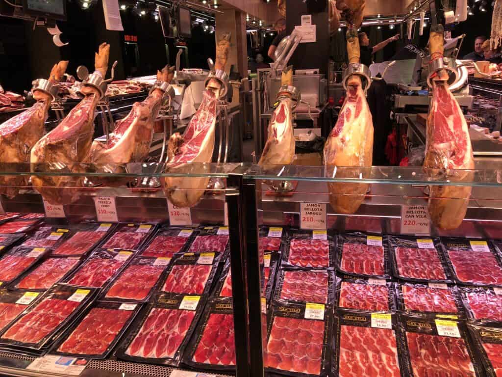 Whole legs of jamón ibérico and bellota on display at a Barcelona market stall with packaged sliced ham — what to eat in Barcelona La Boqueria market