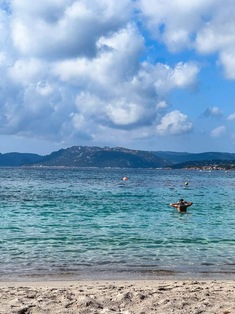 Person swimming in crystal-clear turquoise water at a Corsican beach with mountain island views in the distance — best beaches in Corsica