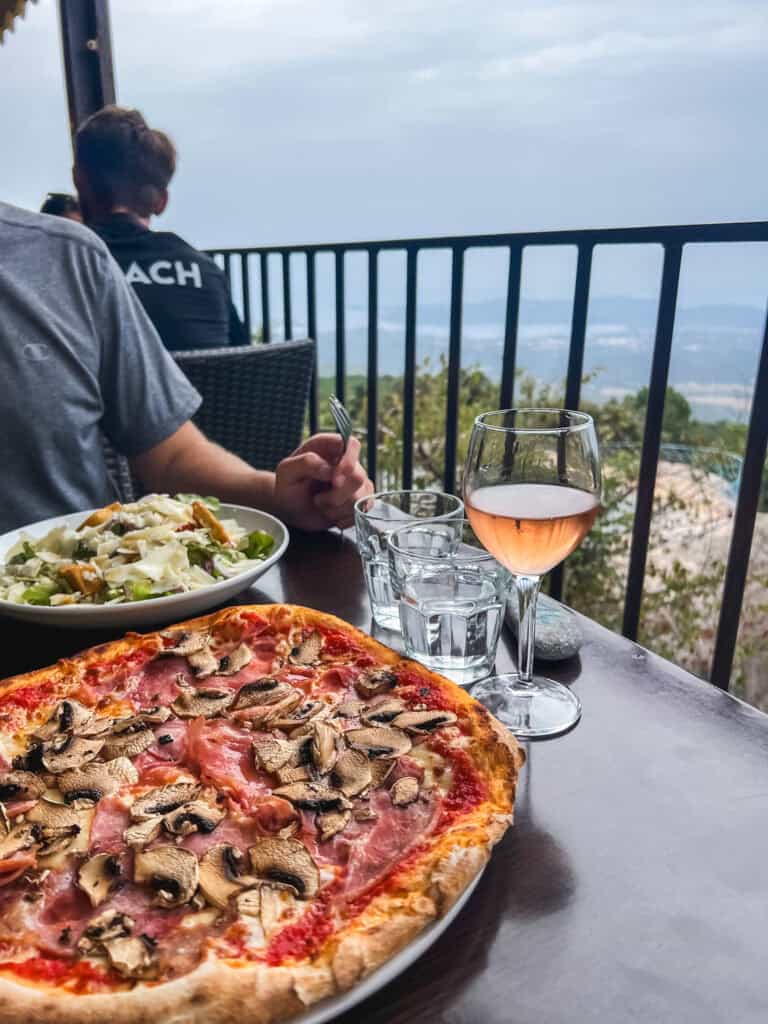 Pizza with ham and mushrooms and a glass of rosé wine on a restaurant terrace with panoramic Corsican mountain and sea views — where to eat in Corsica
