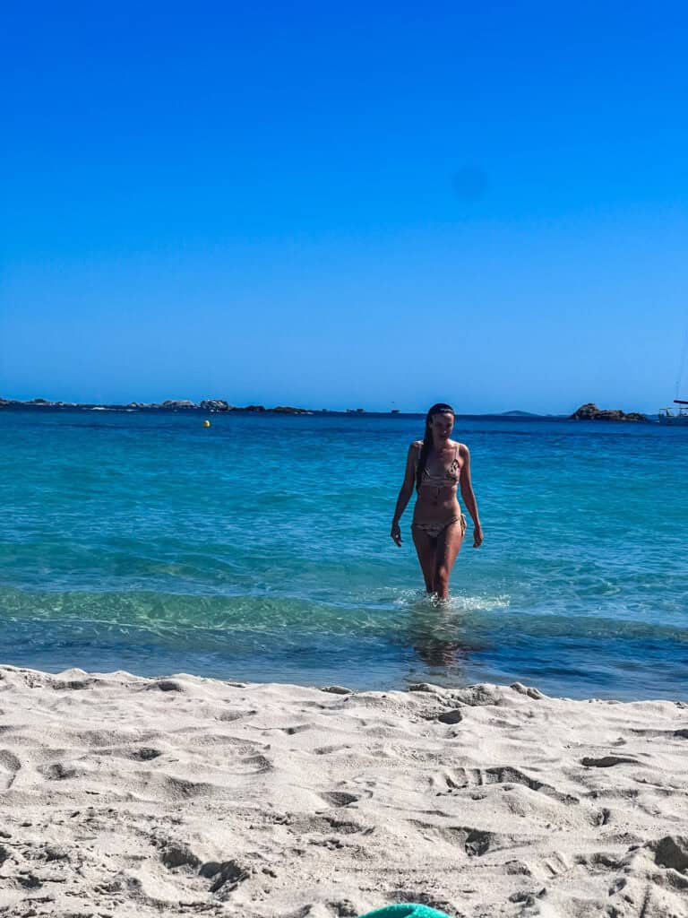 Woman walking out of clear turquoise water onto a white sand beach in Corsica on a sunny summer day — best beaches in Corsica France