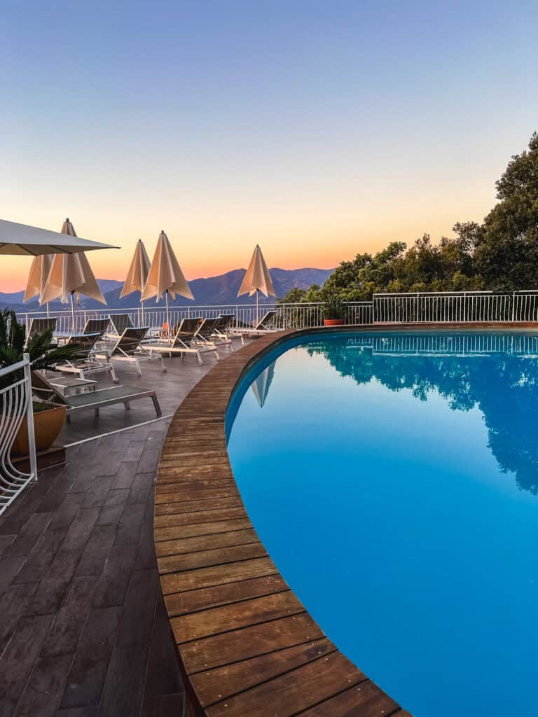 Curved hotel pool at sunset with sun loungers and mountain silhouette in Corsica — luxury Corsica hotel pool at dusk