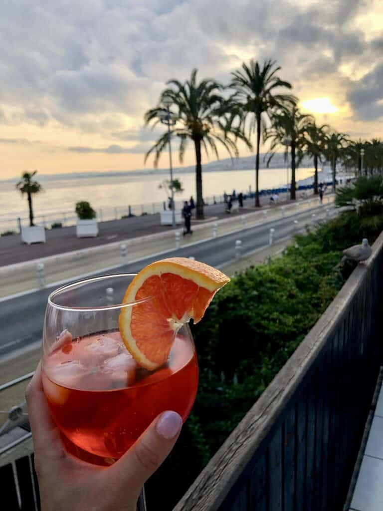 Hand holding an Aperol spritz cocktail on a balcony overlooking the Promenade des Anglais at sunset in Nice — European summer vacations French Riviera
