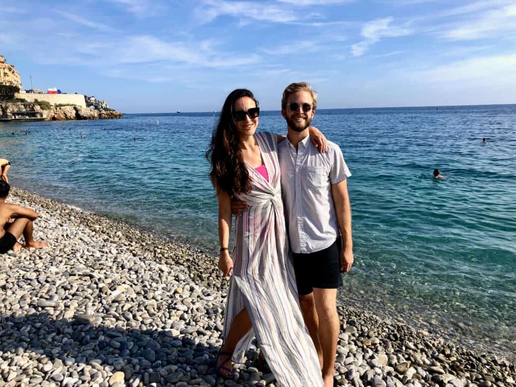 Couple smiling on the pebble beach in Nice with turquoise Mediterranean Sea and the Castle Hill in the background — visiting Nice France summer