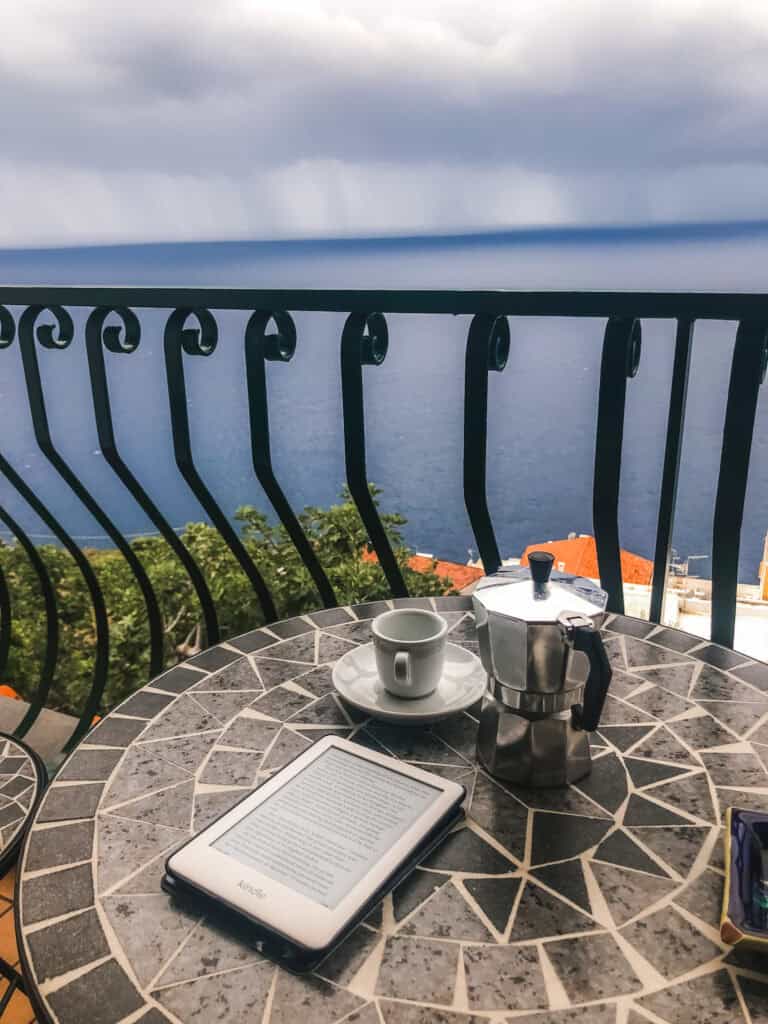Kindle and moka pot espresso on a mosaic balcony table overlooking the Tyrrhenian Sea and Positano rooftops under stormy skies — slow travel Amalfi Coast Italy