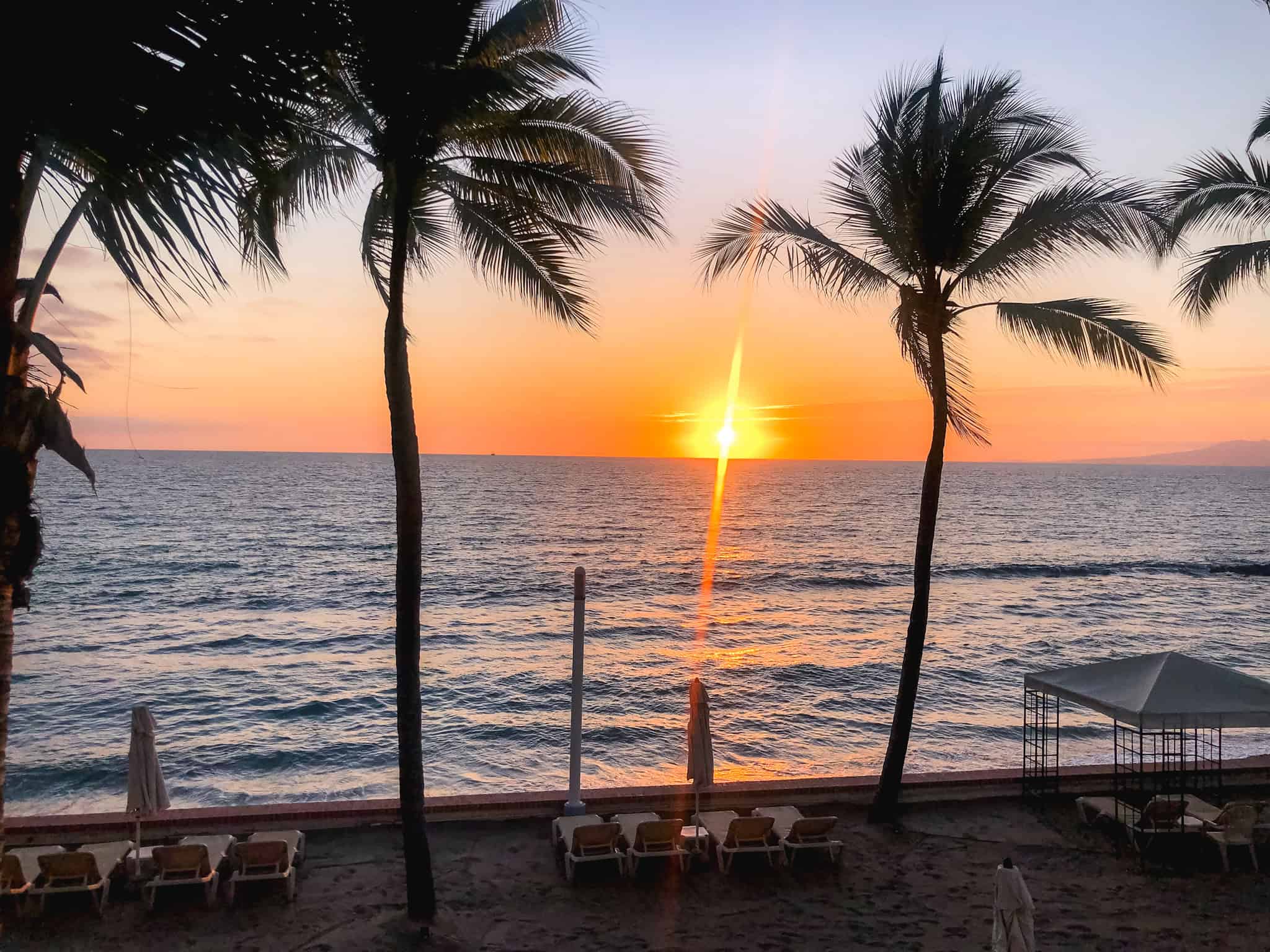 Golden sunset over the Pacific from a resort beach in Puerto Vallarta with palm trees and empty lounge chairs along the shoreline