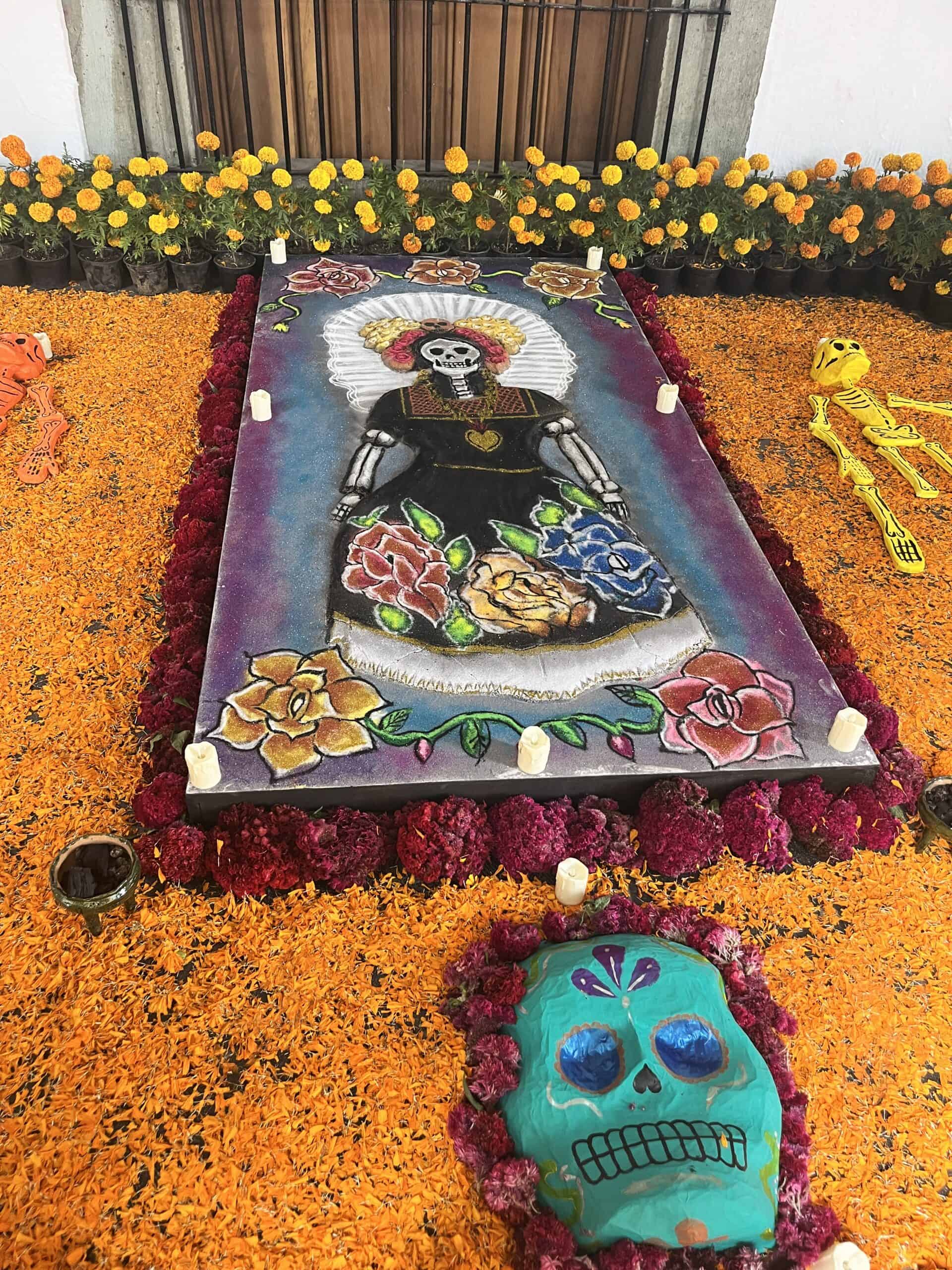 A colorful ofrenda at Mexico City Day of the Dead celebrations featuring a Catrina sand painting surrounded by cempasuchil marigolds, crimson flowers, sugar skull decorations, and flickering candles