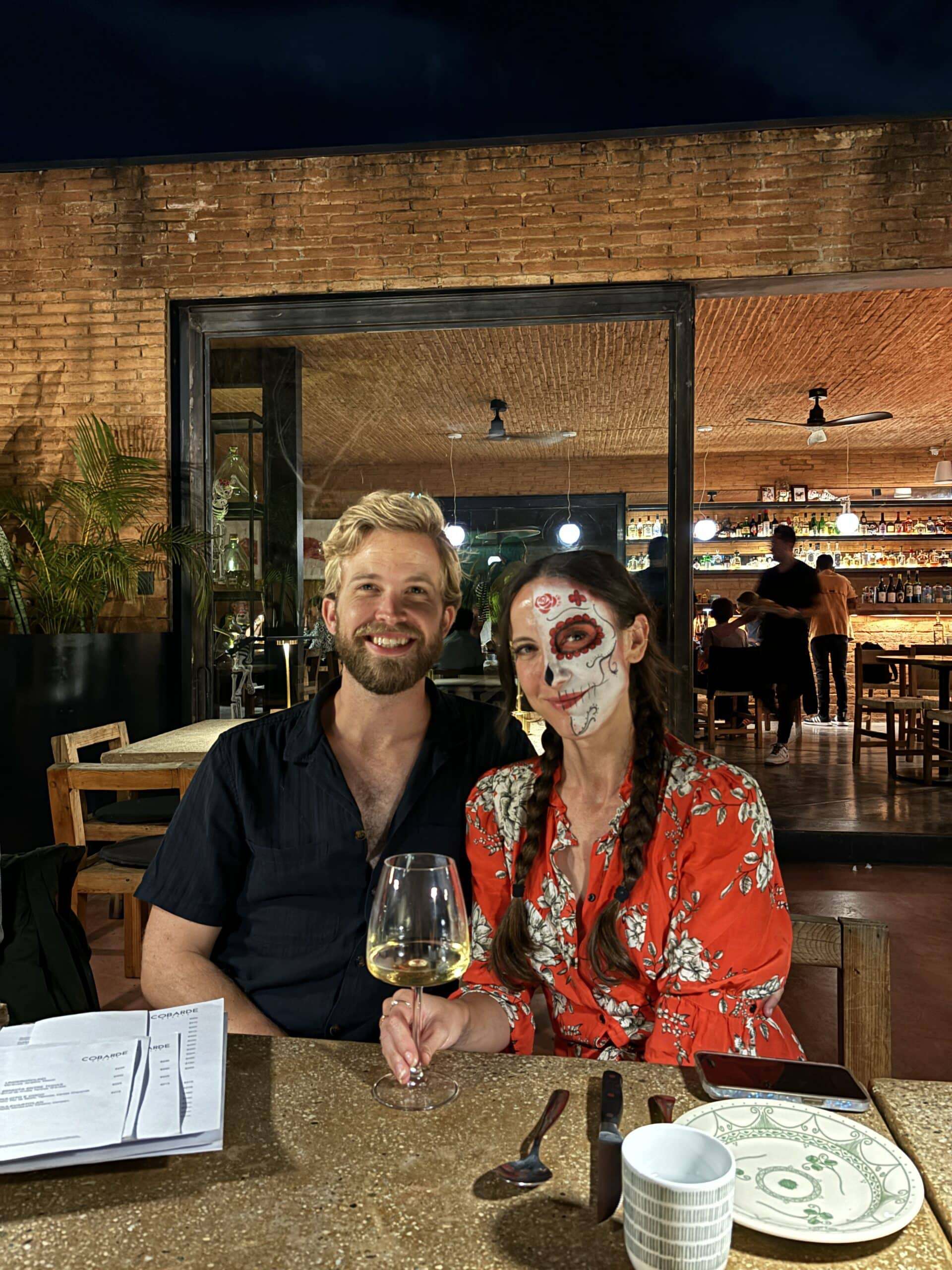 A couple dining at a Mexico City restaurant during Day of the Dead, the woman wearing traditional Catrina sugar skull face paint with a red rose design, sitting at the bar of a warmly lit brick-walled restaurant with a glass of white wine
