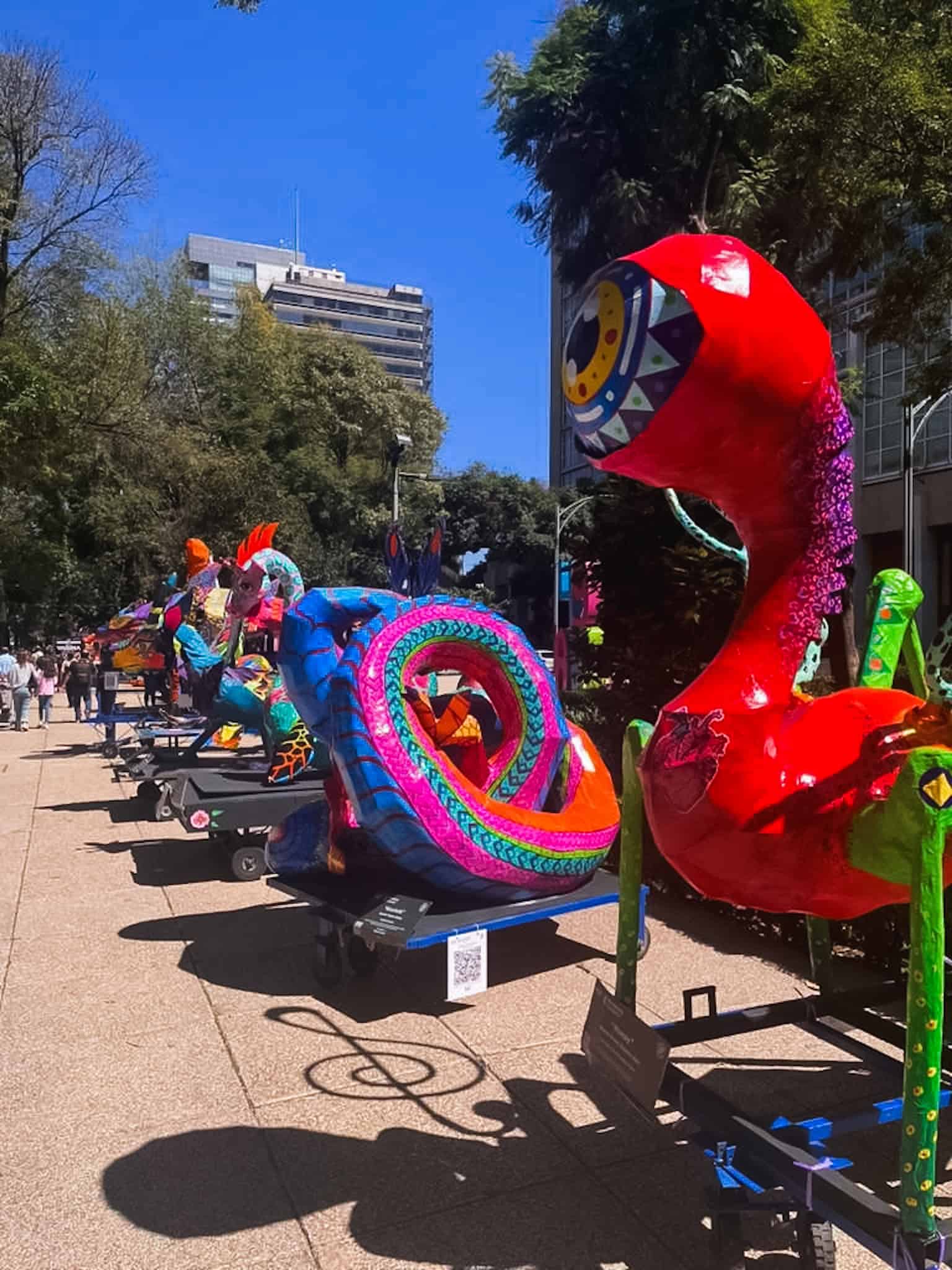 A row of giant alebrije sculptures lining Paseo de la Reforma during CDMX Dia de los Muertos festivities, including a coiled blue serpent and towering red creature with an open jaw