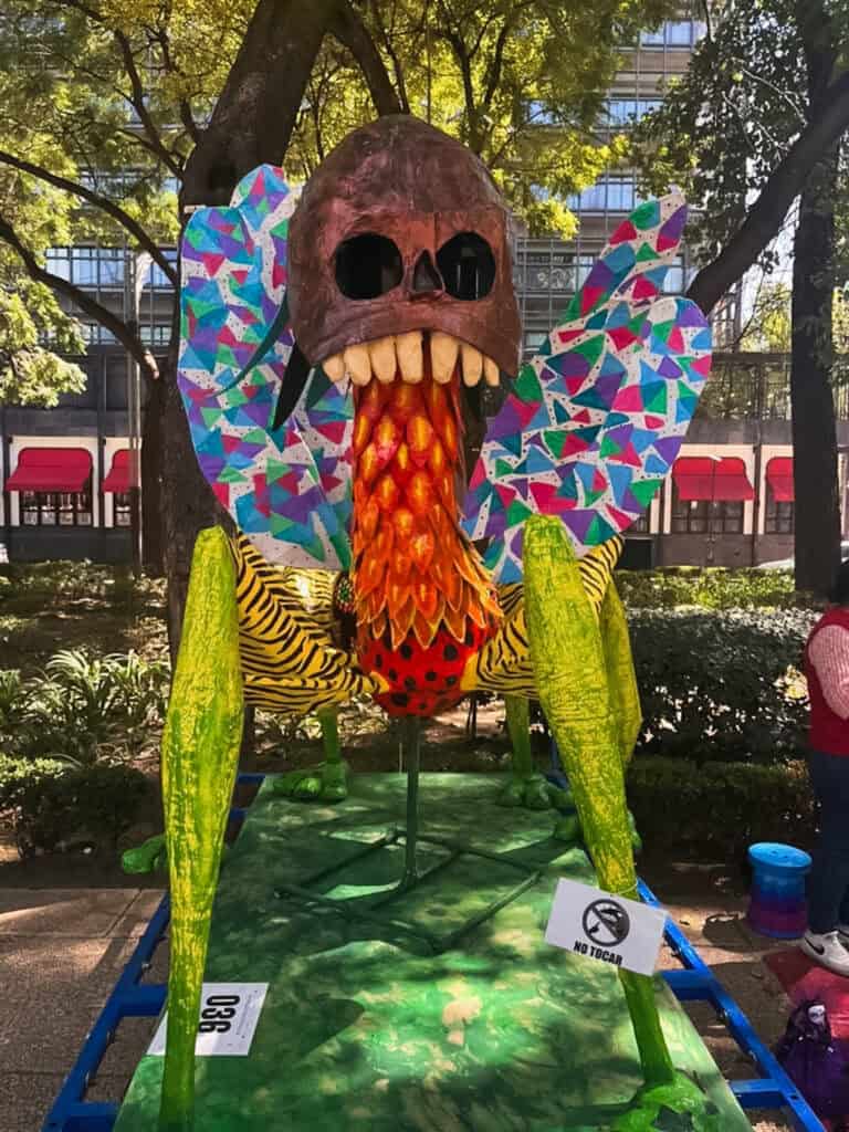 A large papier-mache alebrije creature with a skull head, mosaic wings, and yellow-striped body on display during the Day of the Dead in Mexico City alebrije parade along Reforma