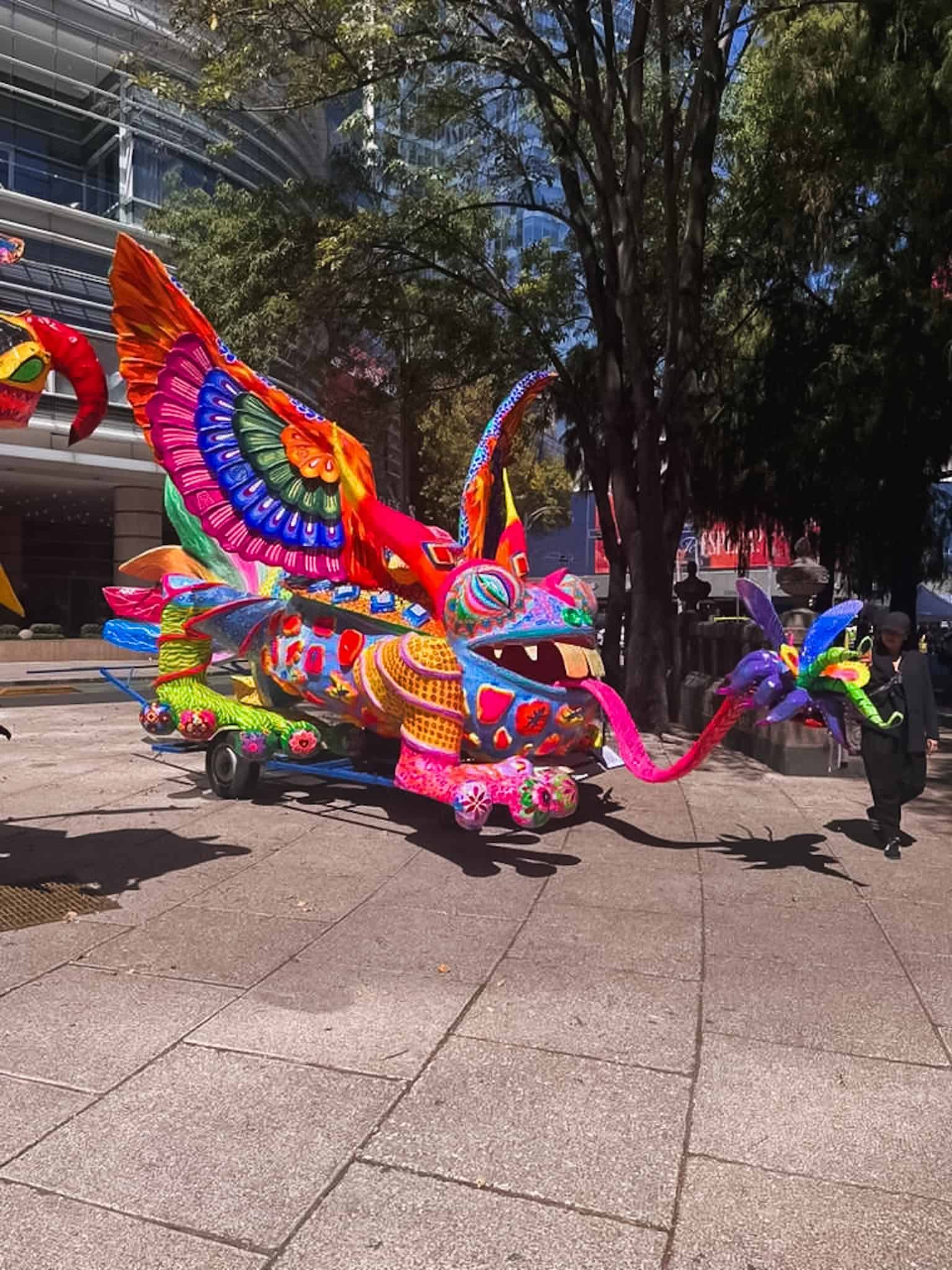 A massive alebrije dragon float with an open mouth and outstretched tongue on Paseo de la Reforma during the Day of the Dead in Mexico City parade, painted in vivid pinks, blues, and greens