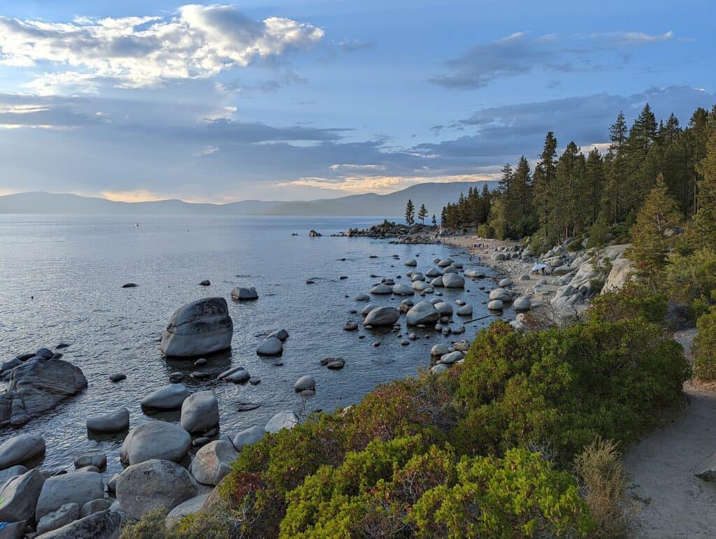 Chimney Beach on the Nevada side of Lake Tahoe, with smooth granite boulders scattered along the shoreline, low scrub brush, tall pines, and a calm blue-grey lake stretching toward distant Sierra Nevada mountains at dusk