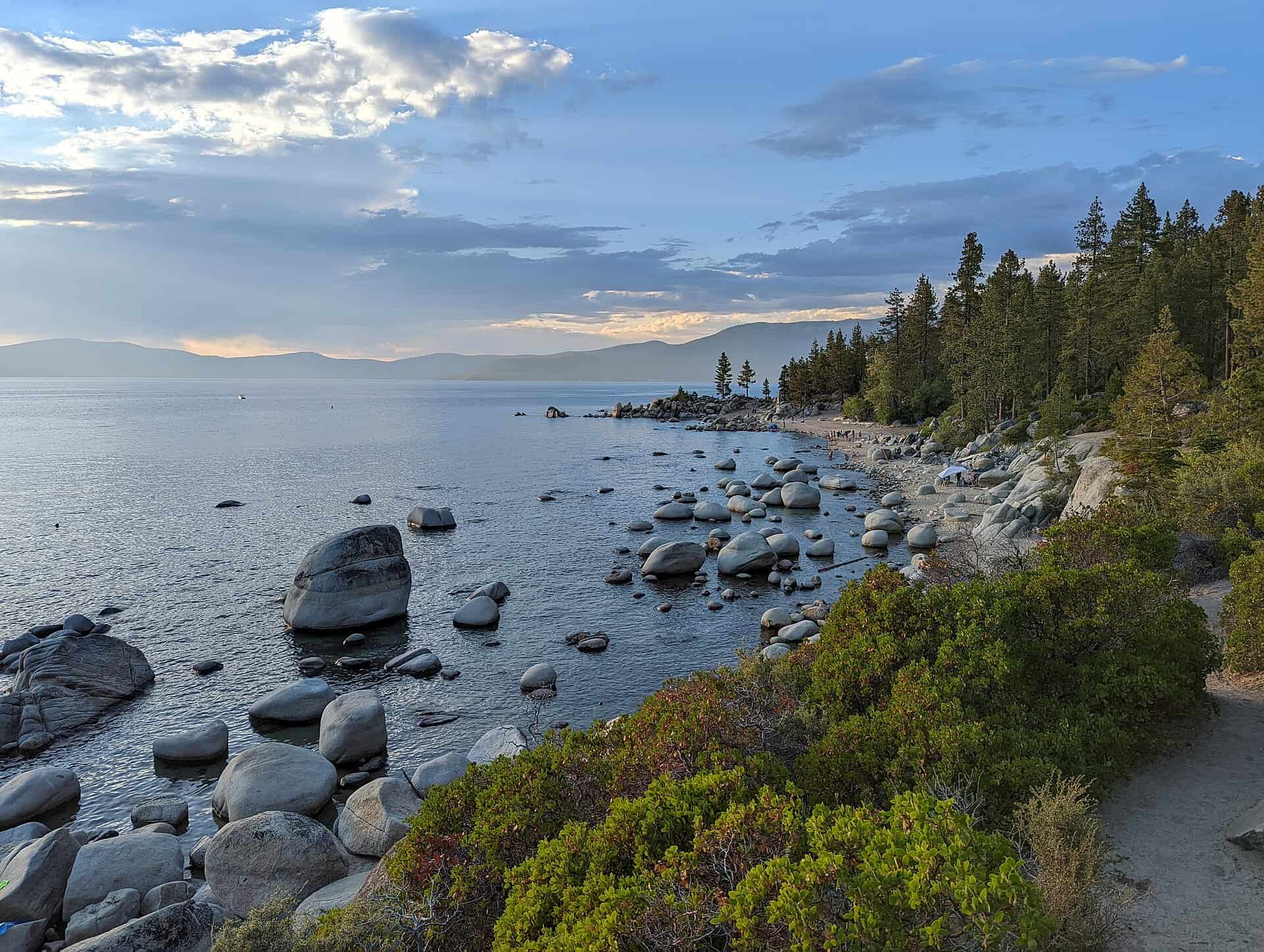 Chimney Beach on the Nevada side of Lake Tahoe, with smooth granite boulders scattered along the shoreline, low scrub brush, tall pines, and a calm blue-grey lake stretching toward distant Sierra Nevada mountains at dusk