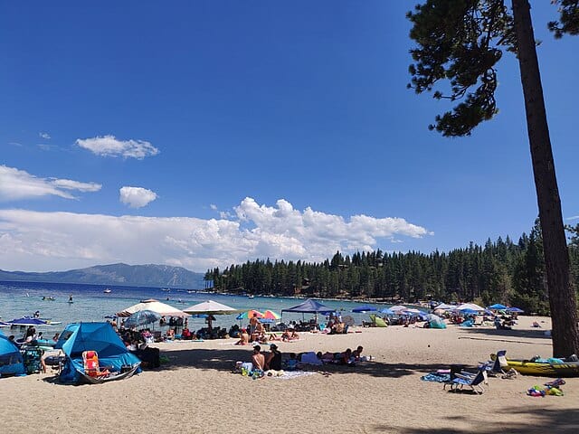 Meeks Bay beach at Lake Tahoe packed with sunbathers under colorful umbrellas and pop-up tents on a hot summer day, with crystal-clear water, pine forest, and mountain ridgeline in the background
