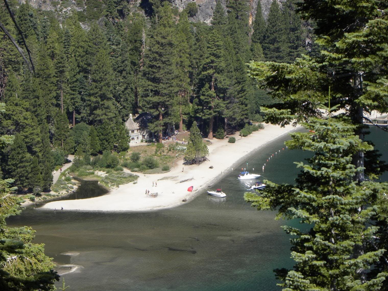 Aerial view of Vikingsholm Beach at Emerald Bay, Lake Tahoe, with the historic Vikingsholm castle visible among the pines, a white sand beach curving along the cove, and two motorboats anchored in the clear green water