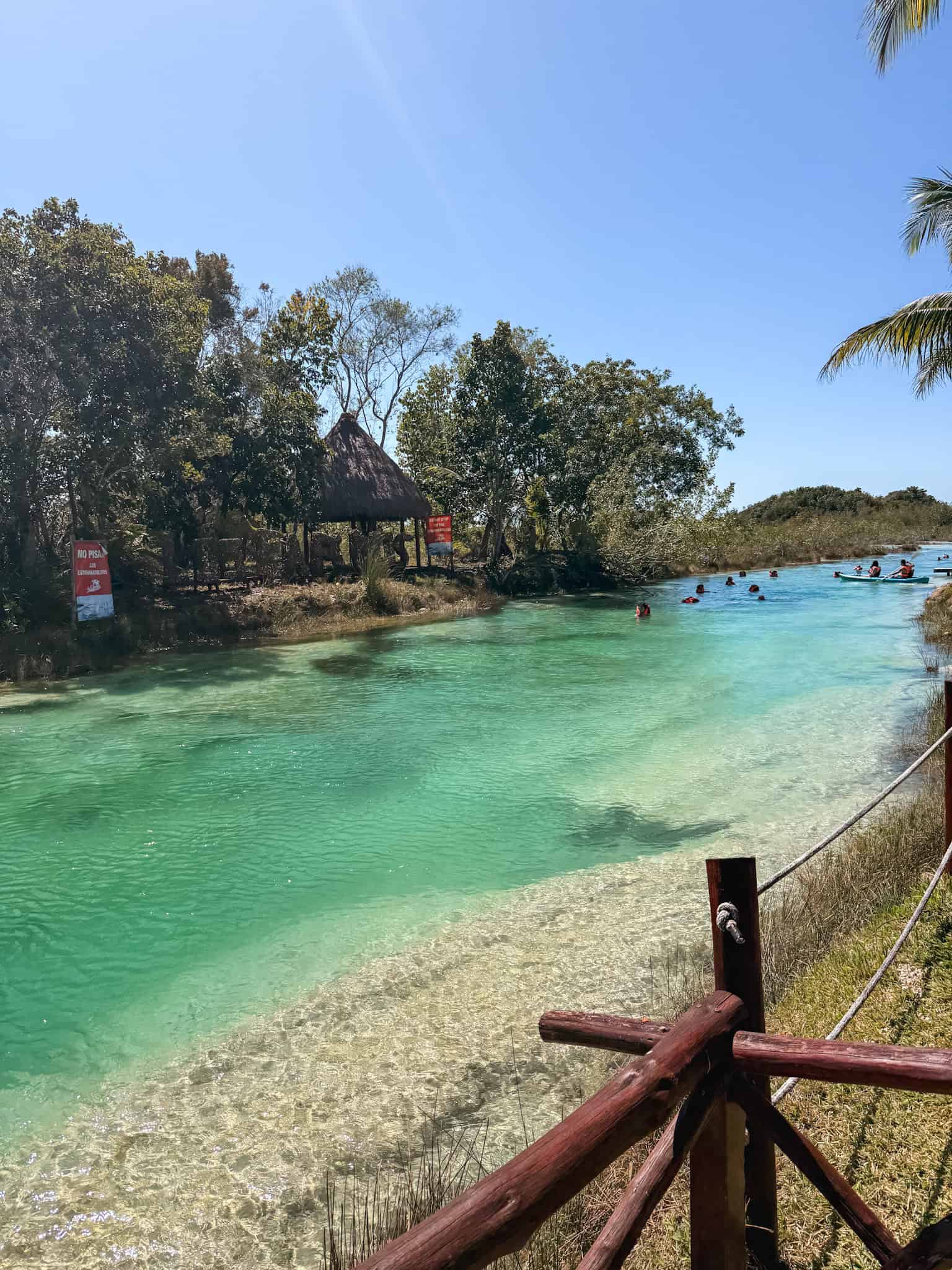 Swimmers and kayakers floating through the emerald green river channel at Los Rapidos Bacalar, with a thatched palapa and wooden fence lining the crystal clear water