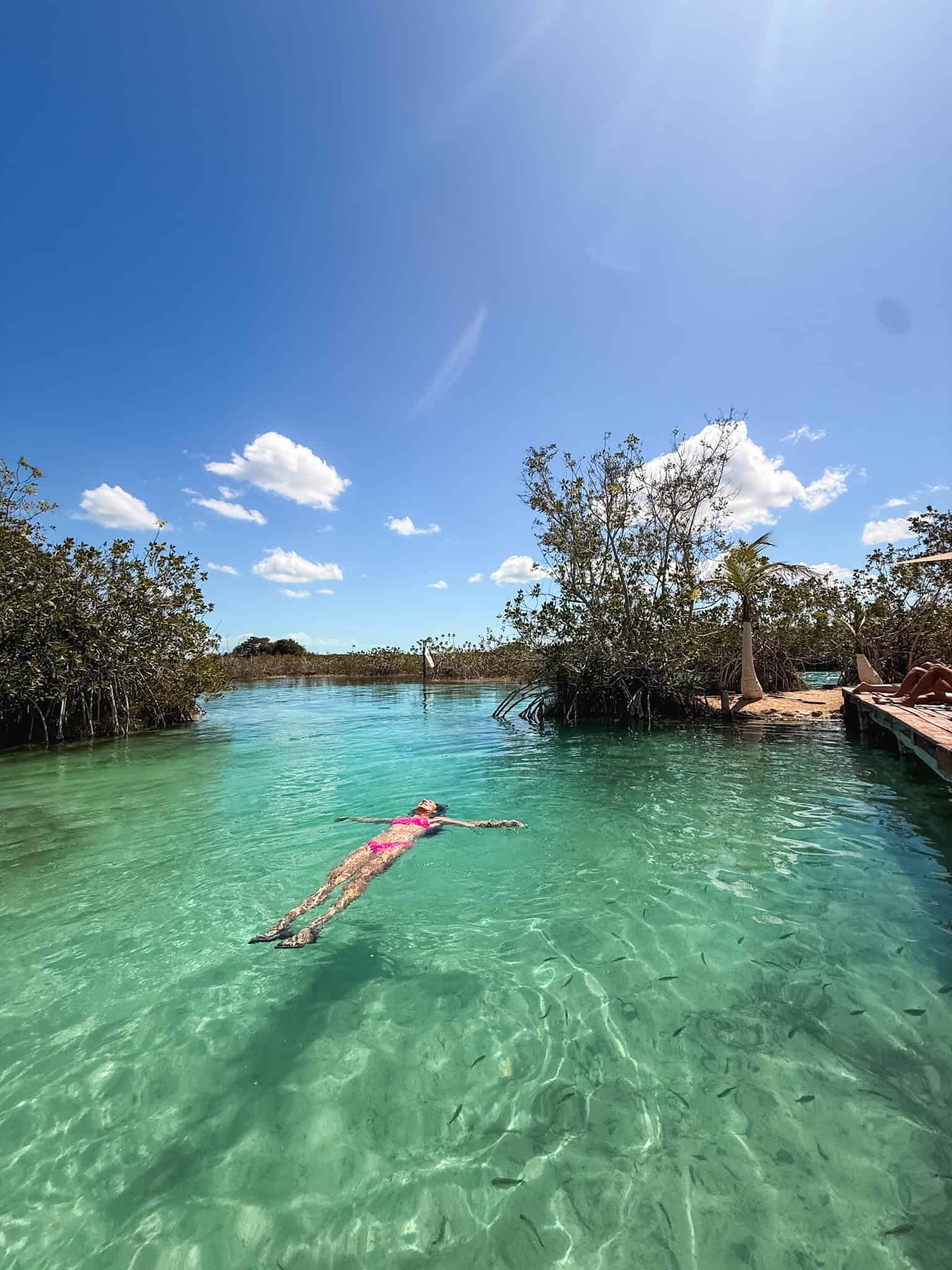 Woman in a pink bikini floating on her back in the crystal clear turquoise water at Los Rapidos Bacalar, with small fish visible beneath the surface and mangroves lining the river channel