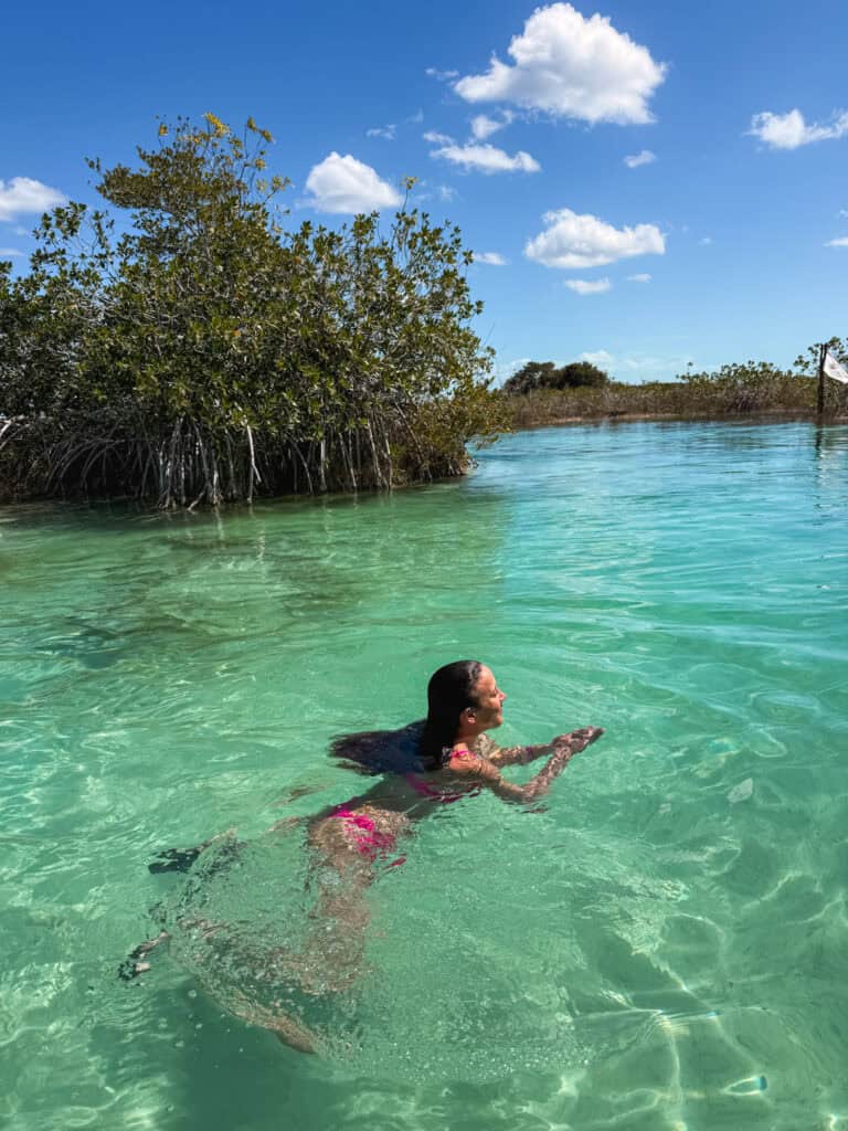 Woman swimming through the shallow turquoise water at a Bacalar lagoon club, surrounded by mangrove trees reflected in the clear green water on a sunny day in Quintana Roo