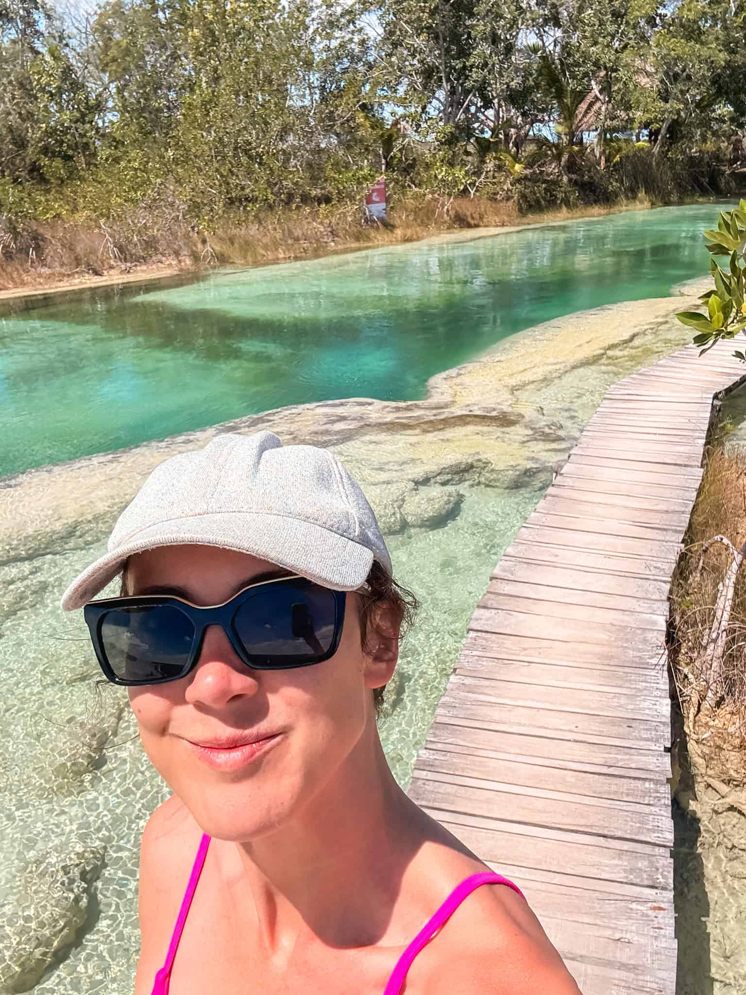 Woman in a pink swimsuit, cap, and sunglasses taking a selfie in the turquoise river at a Bacalar lagoon club, with a wooden boardwalk and mangroves in the background