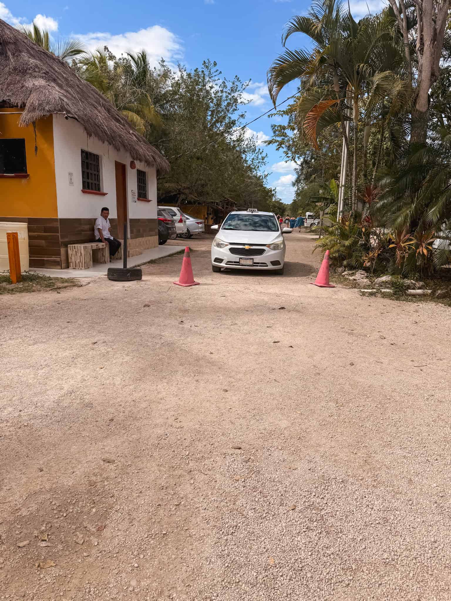 White taxi waiting on a dirt road outside Los Rapidos Bacalar beside a yellow building with a thatched palapa roof, showing how to get to the river club from Bacalar town