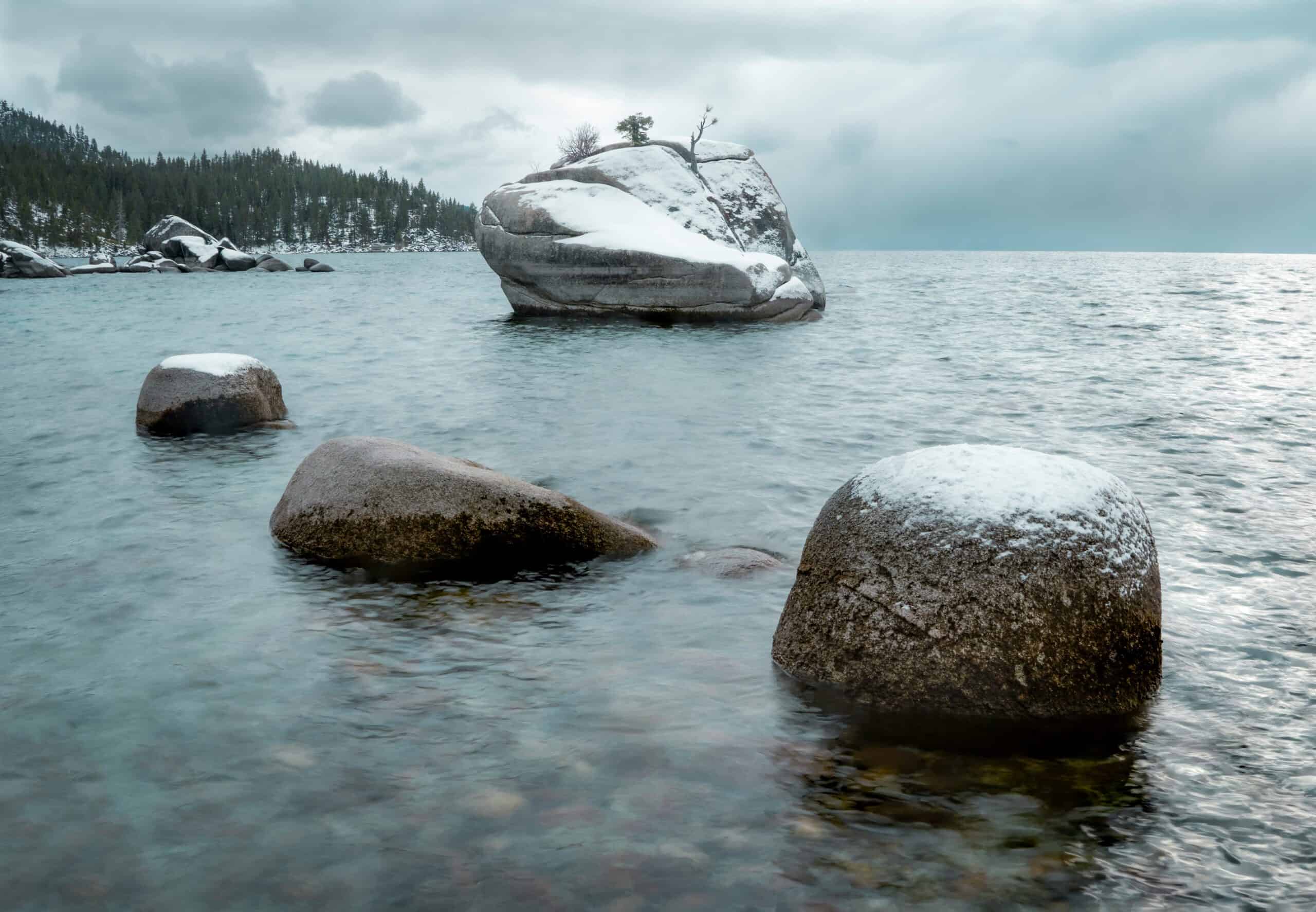 Snow-dusted Bonsai Rock rising from the grey winter water of Lake Tahoe on the Nevada side, with a lone windswept tree clinging to its top