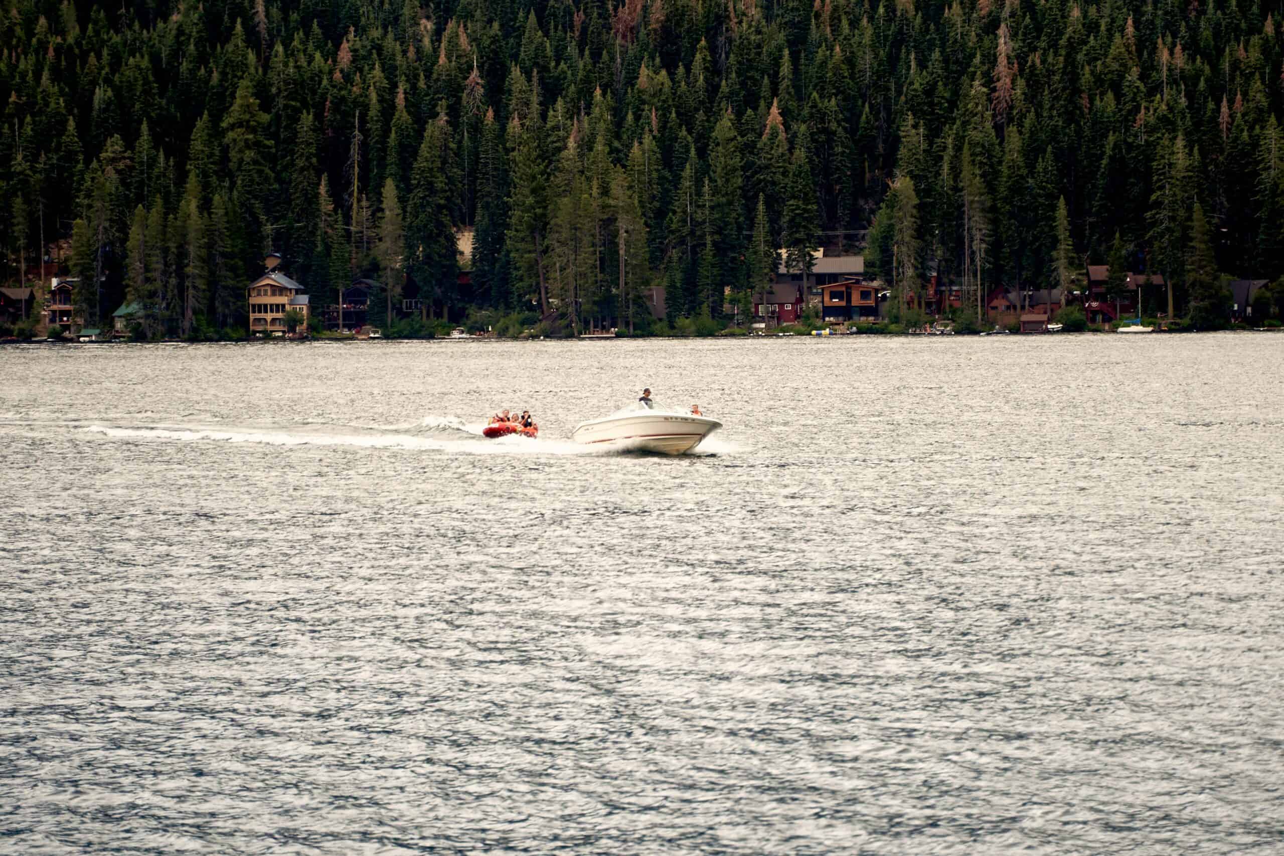 Speedboat towing riders on an inflatable tube across Donner Lake with lakeside cabins and dense pine forest lining the far shore