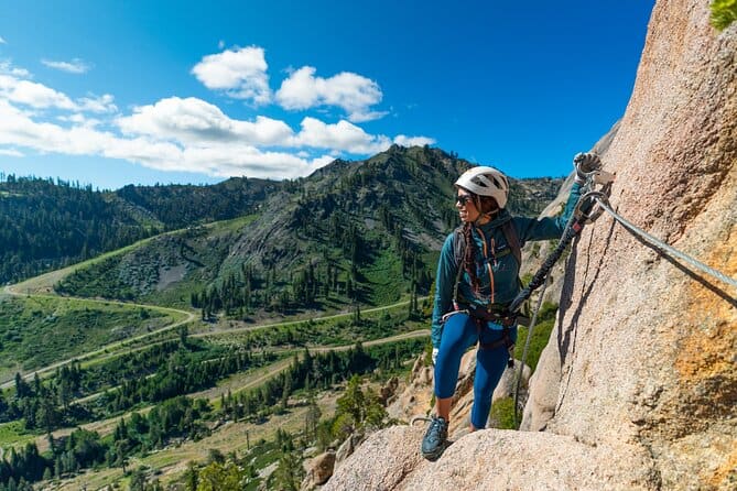 Woman in helmet and harness climbing a via ferrata route above a Sierra Nevada valley near Lake Tahoe, clipped into a steel cable on a granite rock face