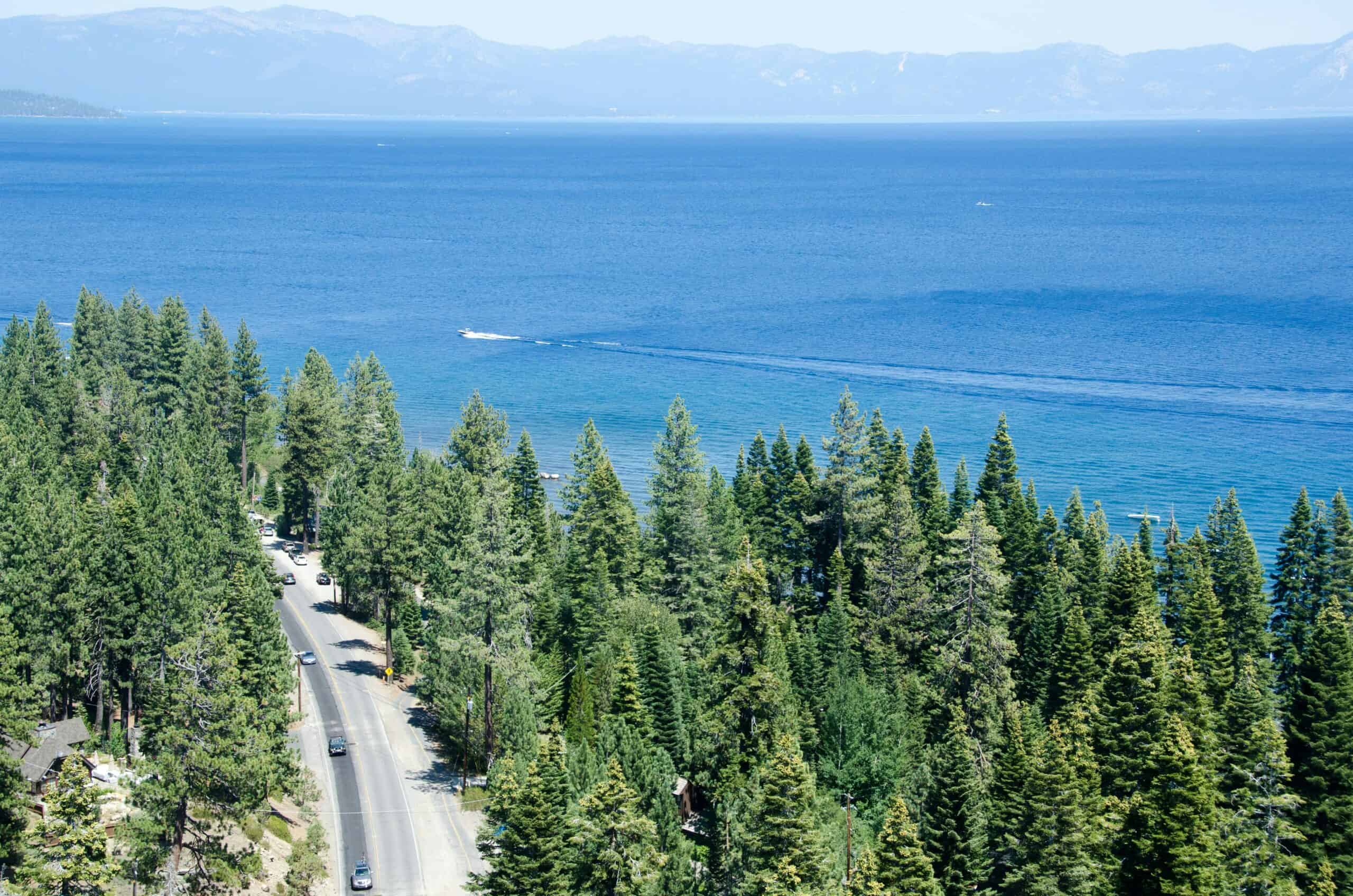 Aerial view of Highway 89 running through tall pine trees along the western shore of Lake Tahoe with deep blue water and distant mountain ridges