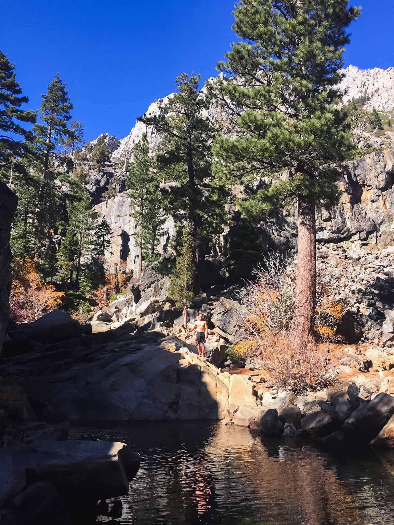 Man standing on granite rocks beside a dark swimming hole surrounded by pine trees and fall foliage below a granite cliff near Lake Tahoe