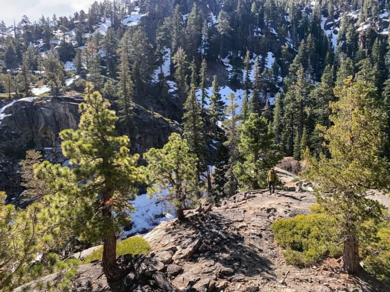 Hiker in a yellow jacket picking a path across a granite ridge above a snow-patched canyon with pines and frozen water below, one of the top things to do in Tahoe in early summer
