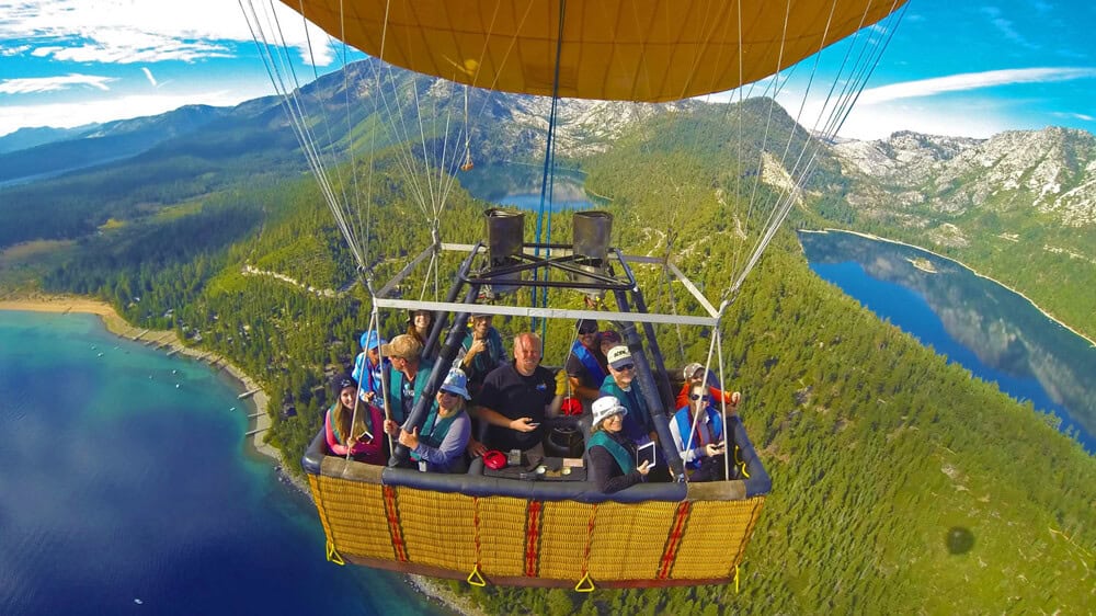 Hot air balloon basket filled with passengers floating above Lake Tahoe with Emerald Bay and the Sierra Nevada visible far below