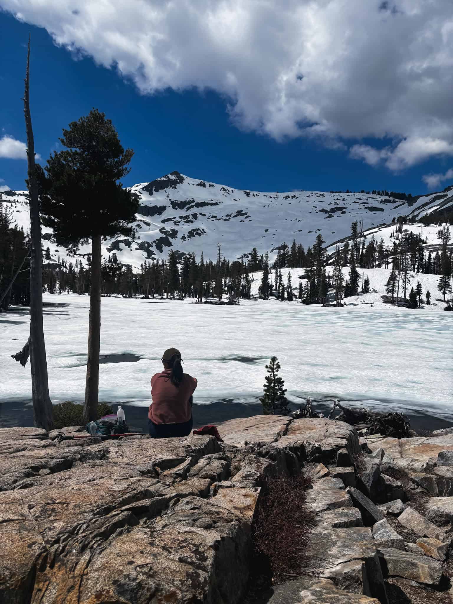 Woman sitting on granite rocks at the edge of a frozen alpine lake below a massive snow-covered peak in the Lake Tahoe backcountry