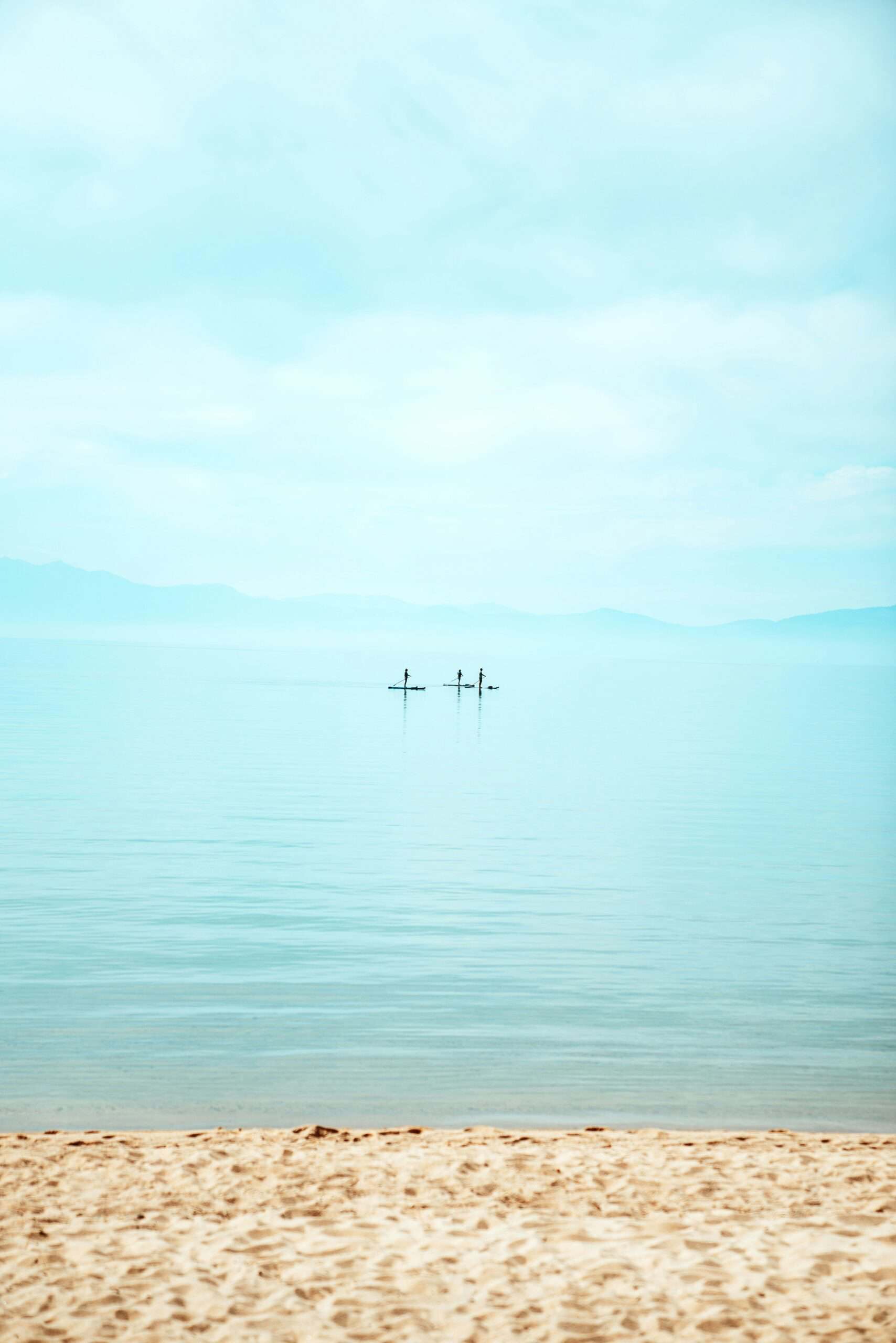Three stand-up paddleboarders silhouetted on the glassy surface of Lake Tahoe seen from a sandy beach, with hazy mountains fading into the pale blue sky