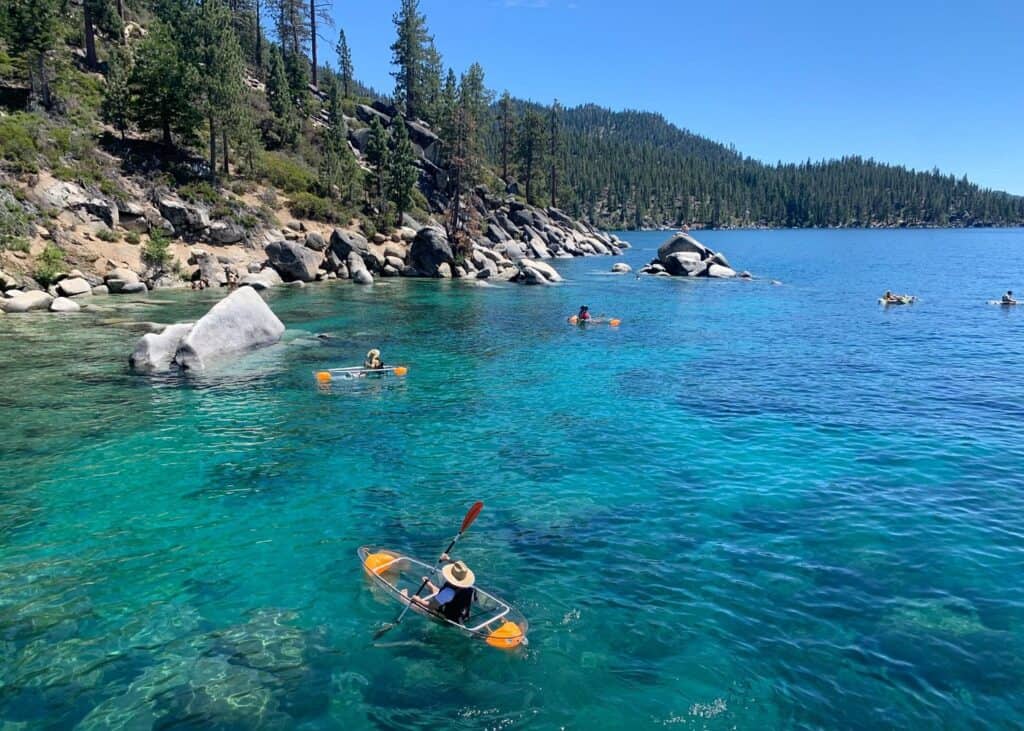 Paddlers in clear-bottom kayaks gliding over turquoise water at Sand Harbor, Lake Tahoe, with granite boulders and a pine-forested shoreline in the background